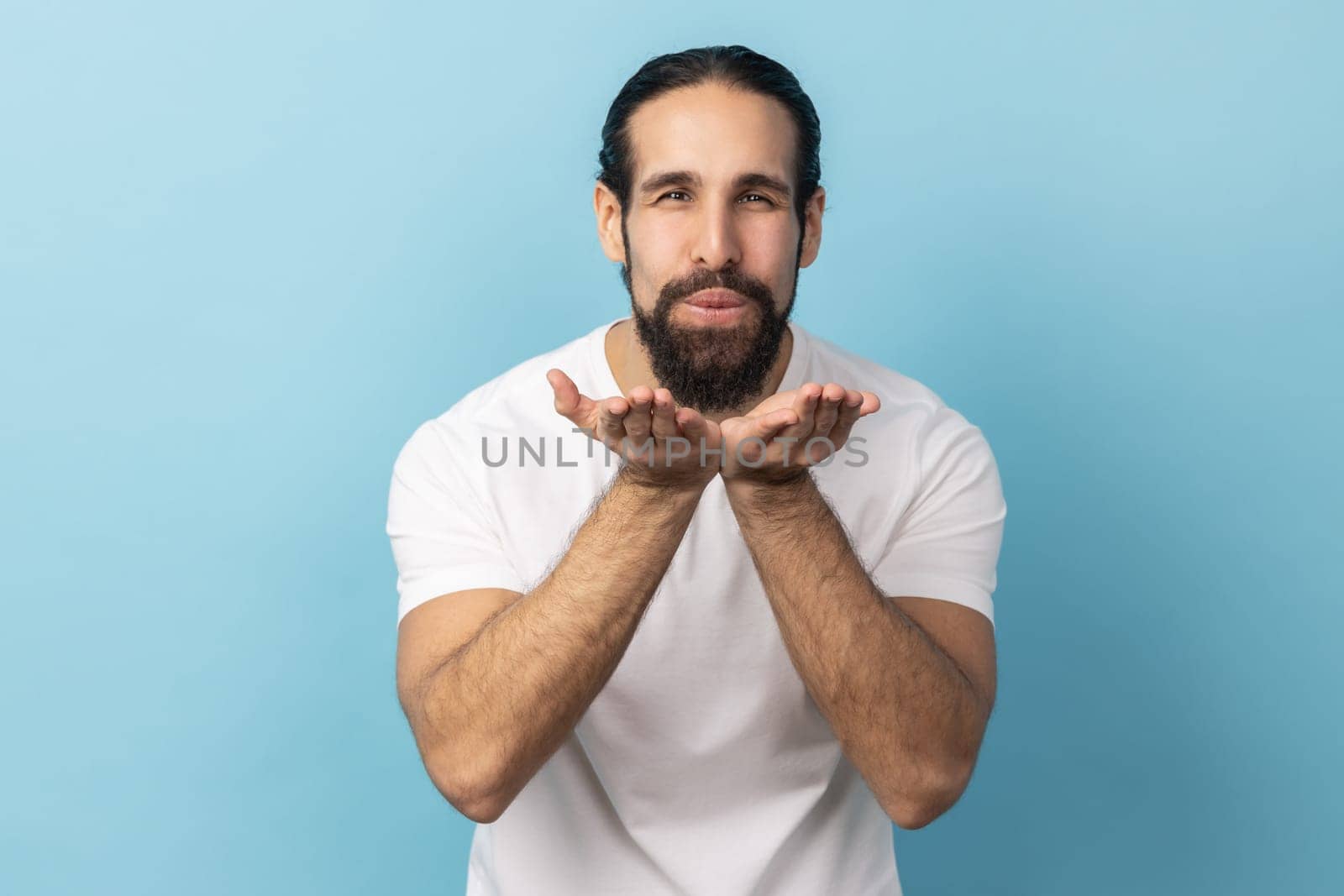 Portrait of man with beard wearing white T-shirt blowing air kiss, folded palm towards mouth, sends mwah coquettish, wants to cheer up his partner. Indoor studio shot isolated on blue background.