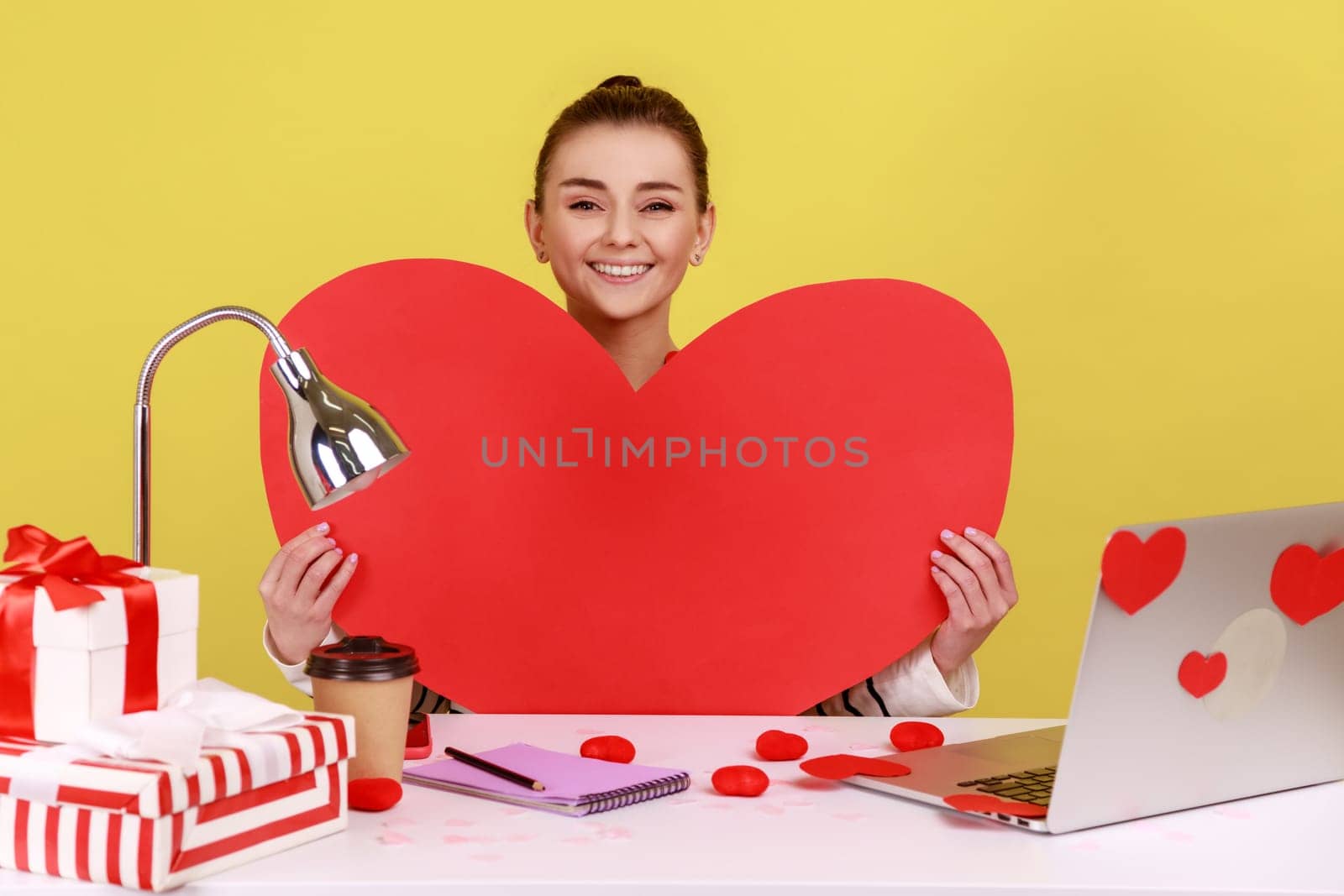 Happy woman holding big red paper heart and smiling, sitting at workplace with laptop in sticky hearts, showing her love. Indoor studio studio shot isolated on yellow background.