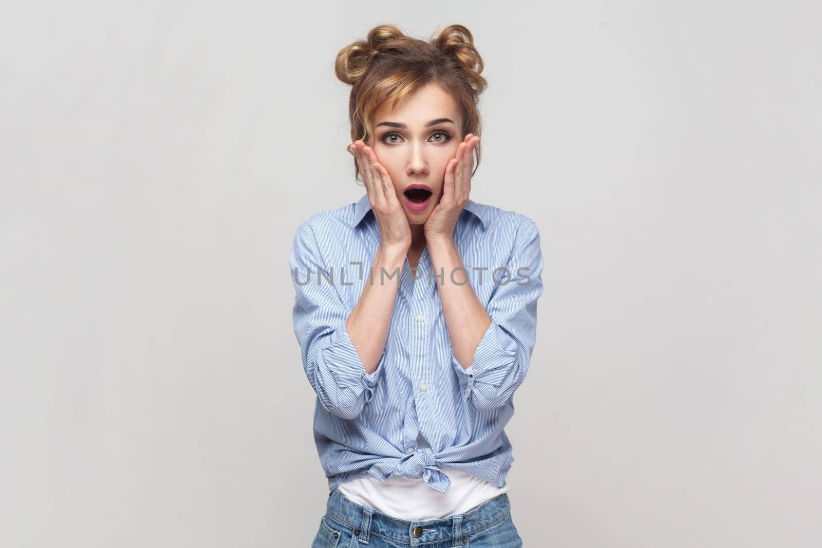 Portrait of embarrassed woman grabs face, having shocked expression, stares with bugged eyes at camera, cannot believe something, wearing blue shirt. Indoor studio shot isolated on gray background.