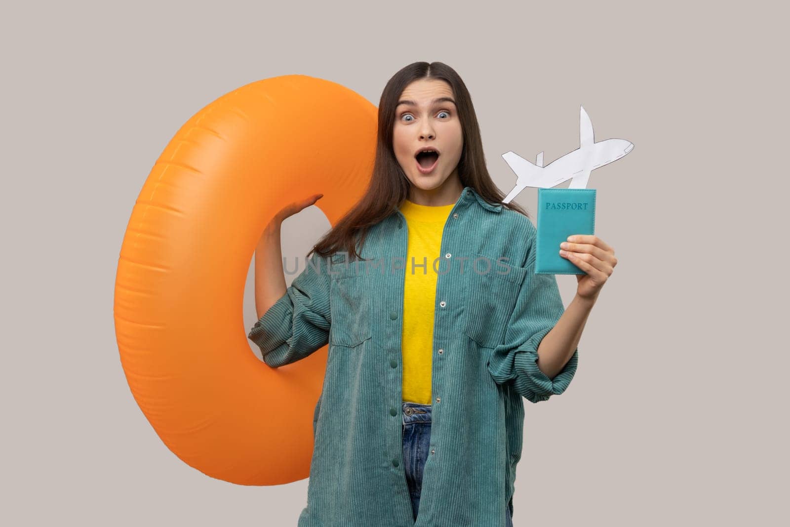 Portrait of amazed shocked woman holding orange rubber ring, passport document and paper airplane, traveling abroad, wearing casual style jacket. Indoor studio shot isolated on gray background.