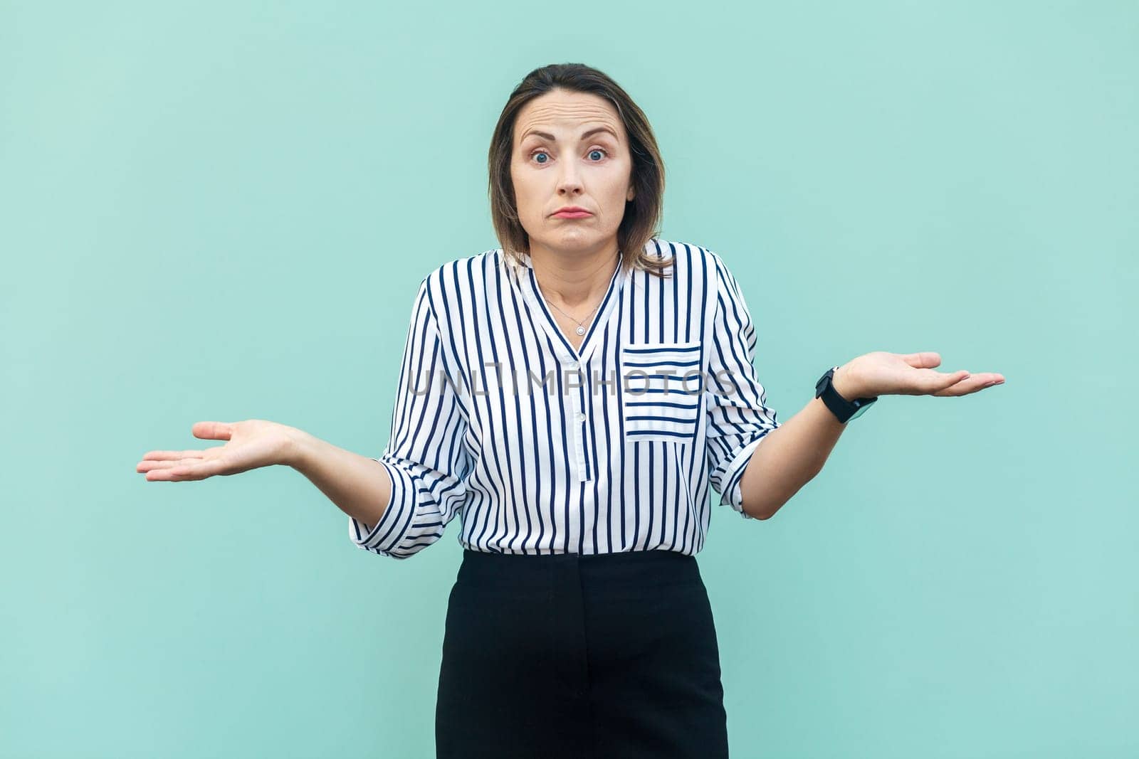 Uncertain clueless attractive middle aged woman wearing striped shirt doesn't know correct answer, looking at camera, shrugging shoulders. Indoor studio shot isolated on light blue background.