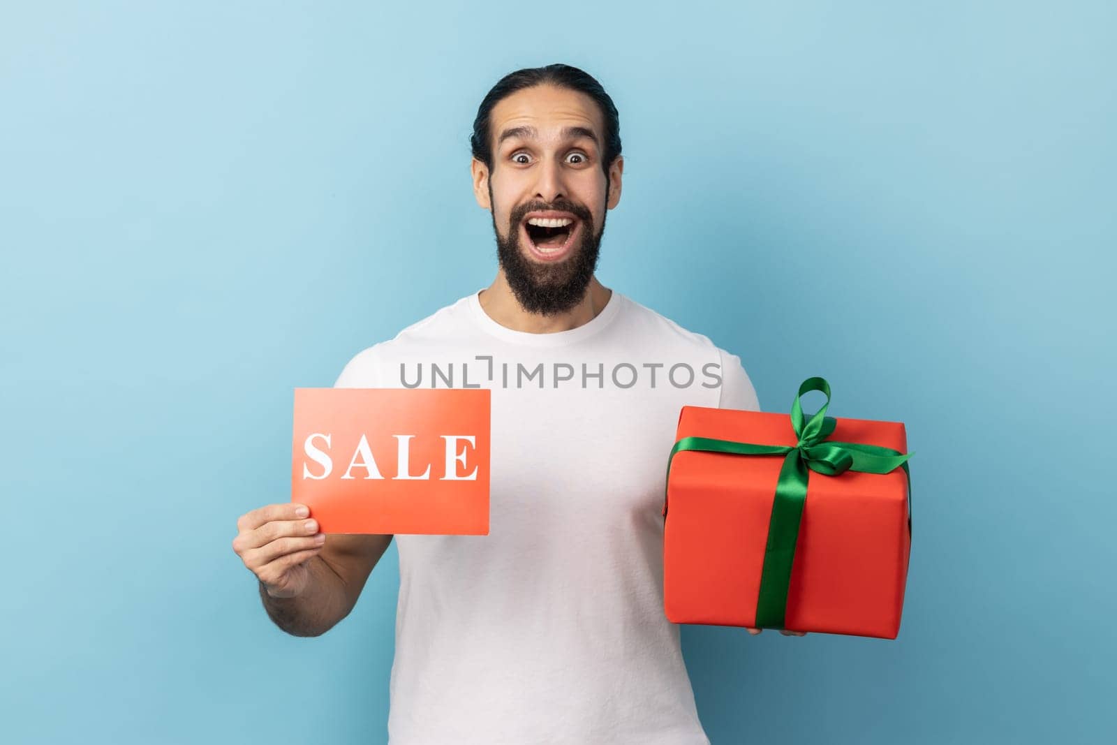 Portrait of amazed surprised man with beard wearing white T-shirt holding red gift box with green ribbon and showing sale coupon, huge sales. Indoor studio shot isolated on blue background.