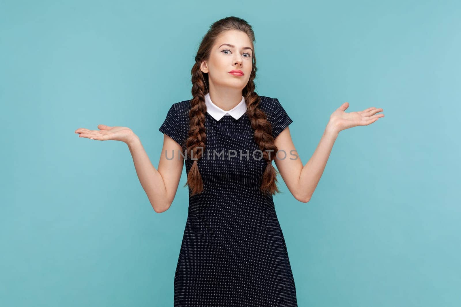 Puzzled uncertain young woman with braids spreads hands aside, shrugging shoulders, don't know how to make a decision, wearing black dress. woman Indoor studio shot isolated on blue background.