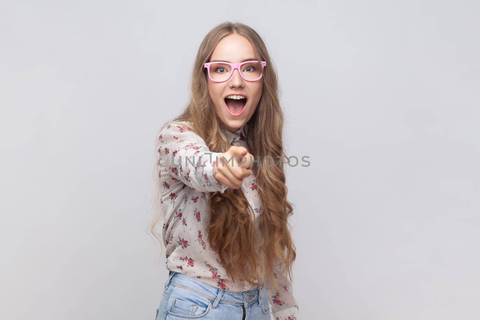 Portrait of surprised woman in glasses with wavy blond hair looking with big eyes and open mouth, pointing finger at camera, suspecting you. Indoor studio shot isolated on gray background.
