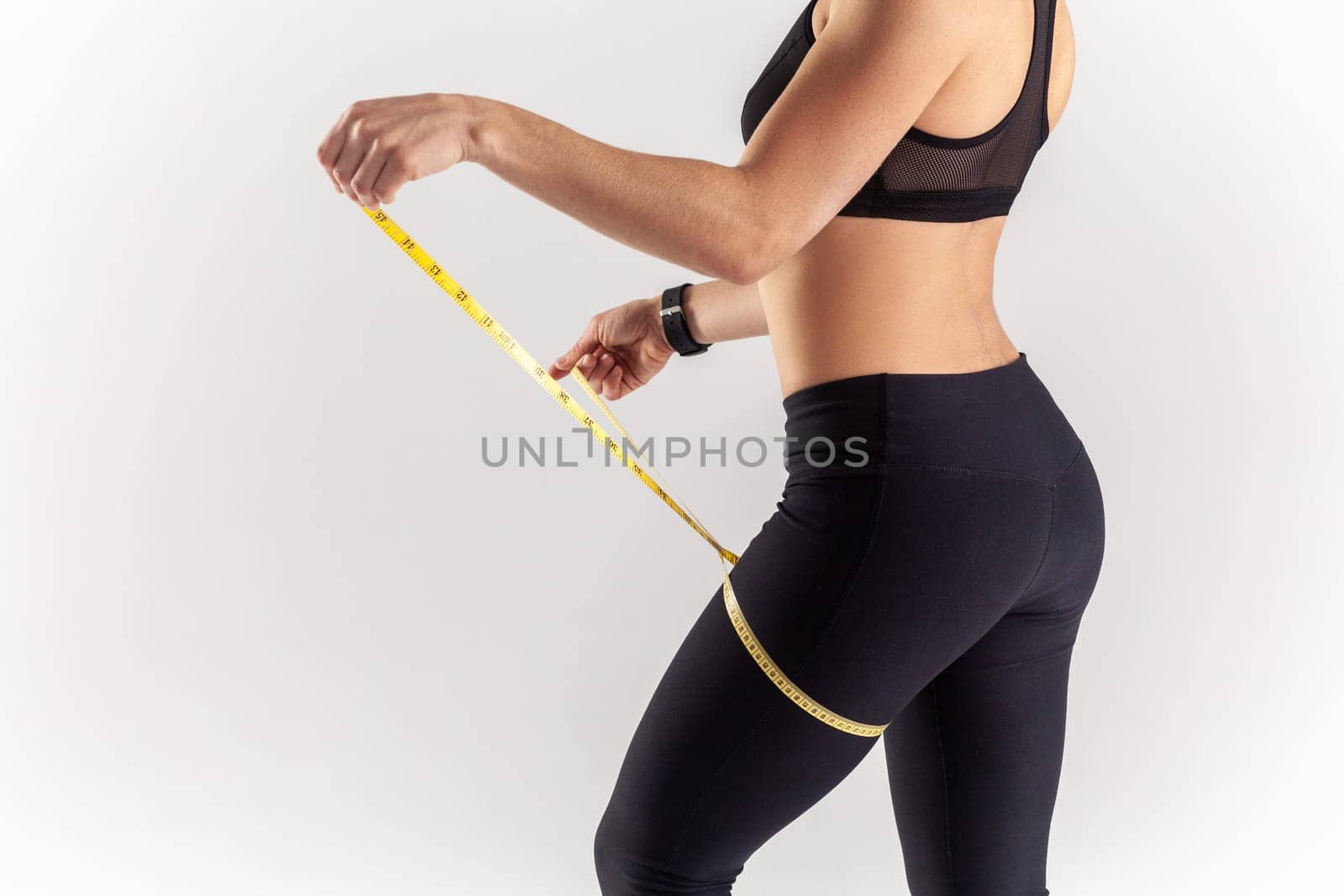 Side view portrait of unknown sporty blonde woman holding centimeter on her leg, checking result after sport marathon, wearing black fitness clothing. Indoor studio shot isolated on gray background.