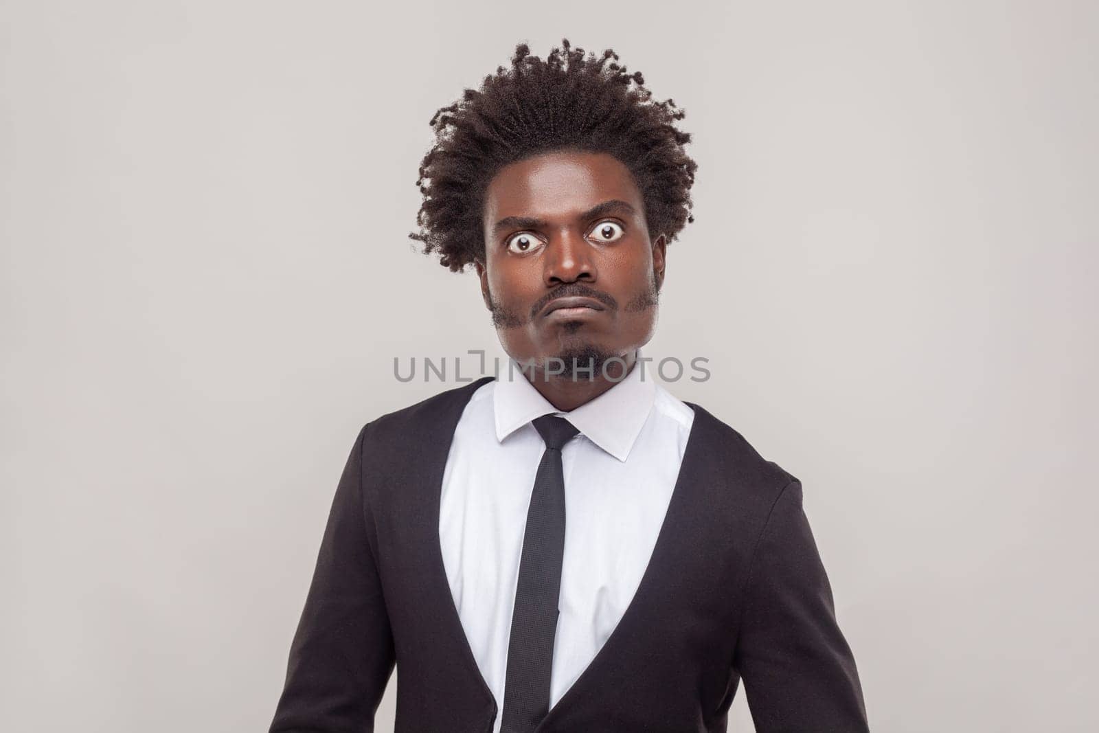 Portrait of man with Afro hairstyle looks with angry strict expression gazes directly at camera, has negative emotions, wearing white shirt and tuxedo. Indoor studio shot isolated on gray background.