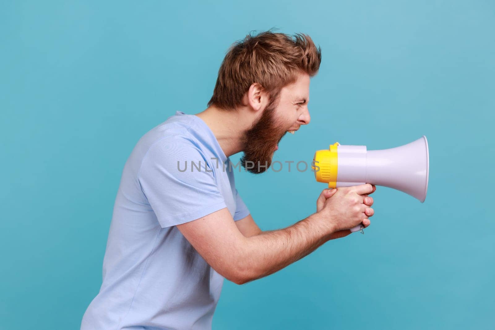 Side view of bearded man holding megaphone near mouth, loudly speaking, screaming, making announcement, paying attention at social problems. Indoor studio shot isolated on blue background.
