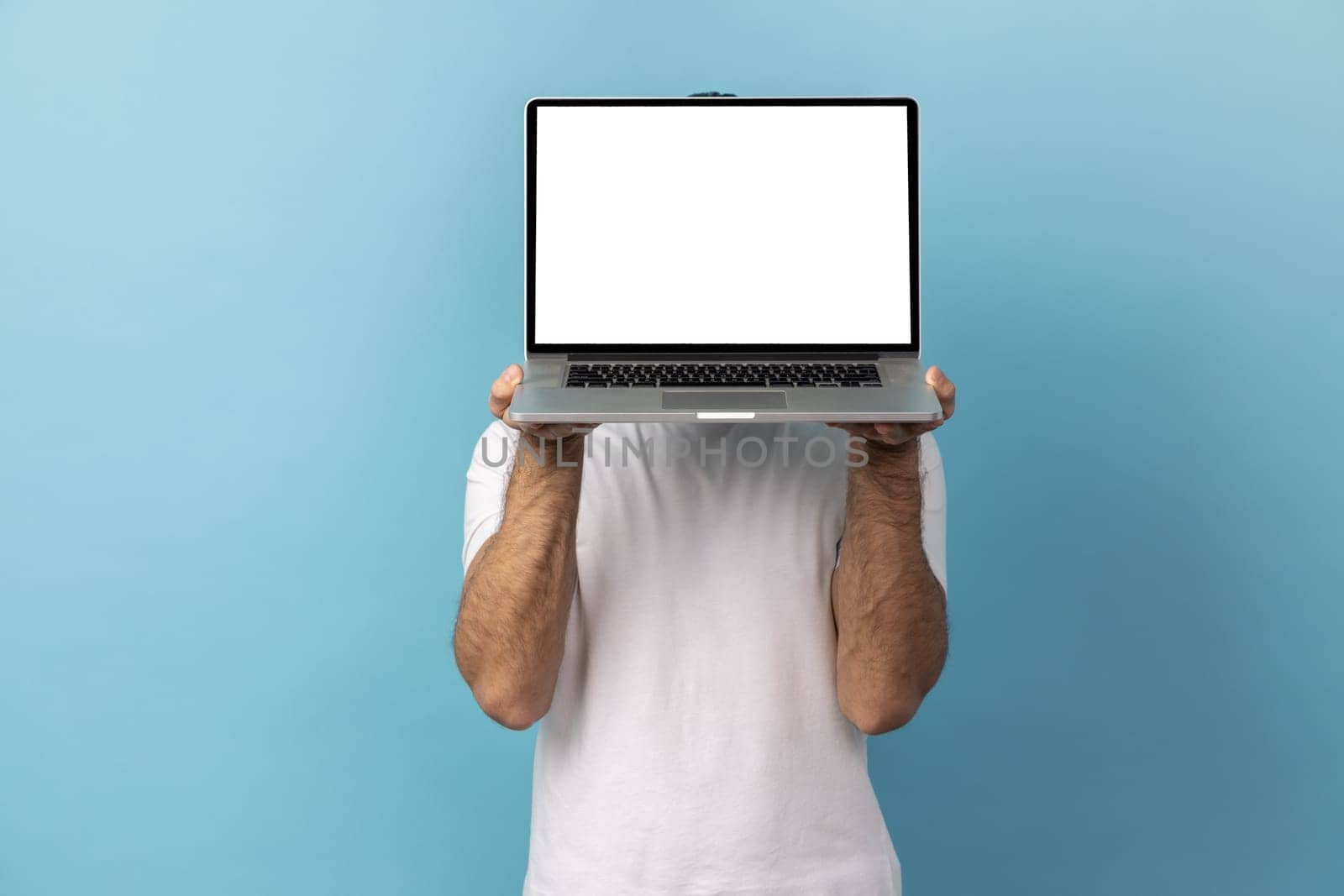 Portrait of man with beard wearing white T-shirt hiding his face behind laptop with blank display for advertisement or promotional text. Indoor studio shot isolated on blue background.