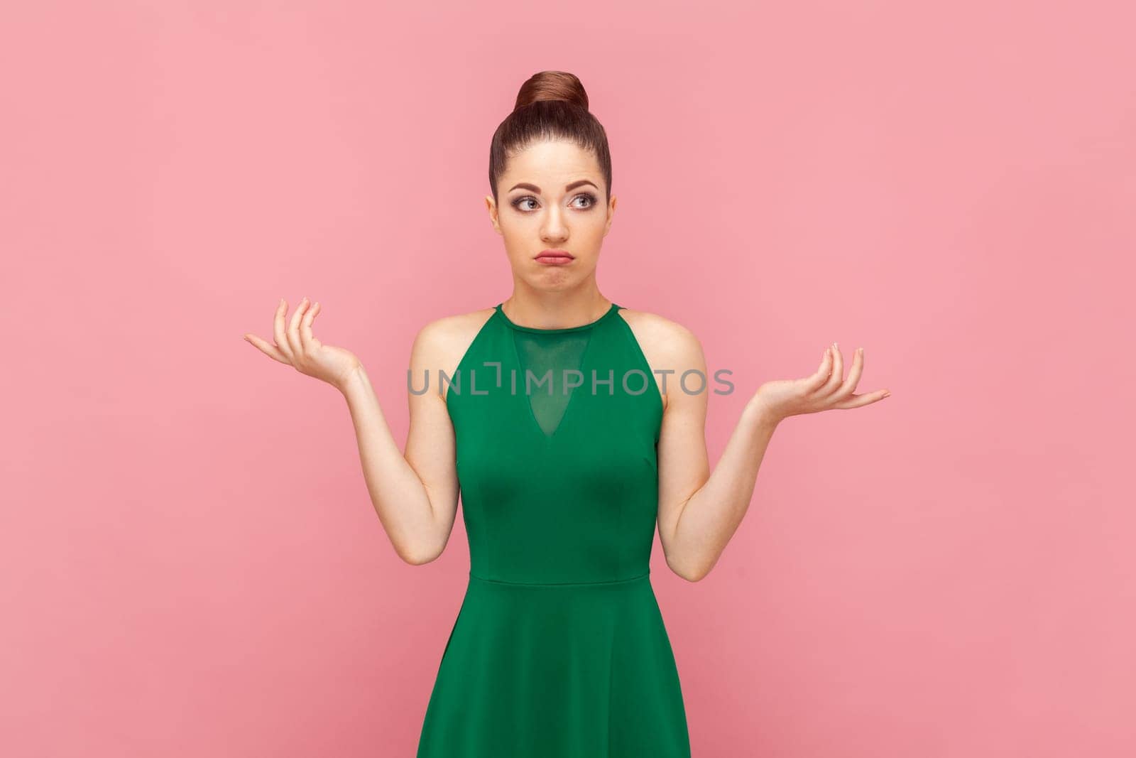 Portrait of confused unsure woman with bun hairstyle standing with spread hands, looking at camera with uncertain expression, wearing green dress. Indoor studio shot isolated on pink background.