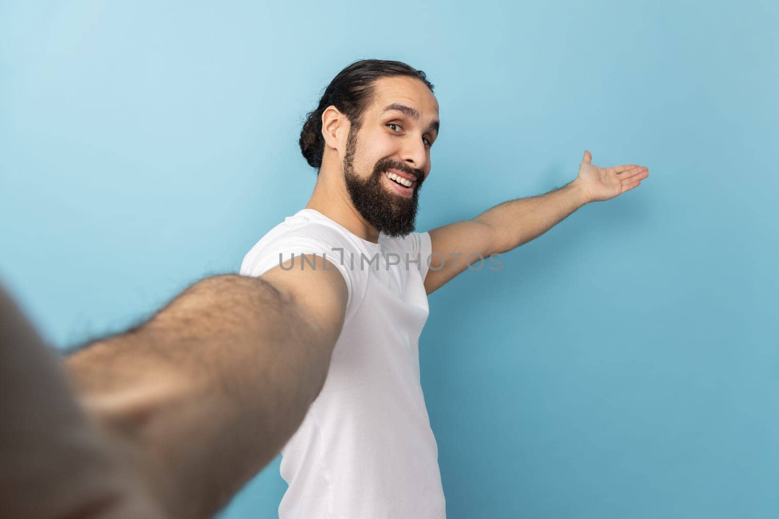Portrait of king friendly man with beard wearing white T-shirt looking at camera with smile, making welcome gesture, taking selfie POV. Indoor studio shot isolated on blue background.
