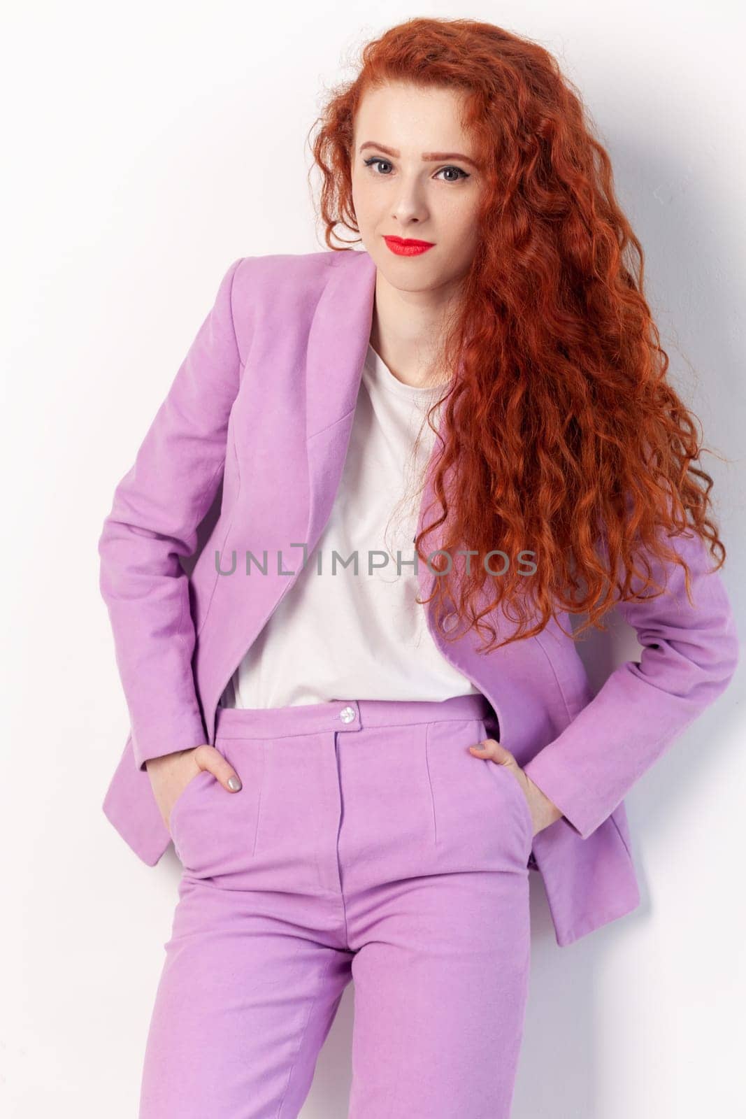 Portrait of self-confident attractive beautiful red haired woman with wavy hair, standing with hands in pockets, looking at camera, wearing lilac suit. Indoor studio shot isolated on gray background