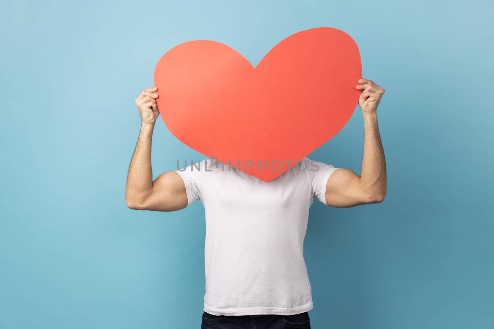 Portrait of man wearing white T-shirt hiding behind big red paper heart, holding symbol of love care, cardiac issues, demonstrating affection. Indoor studio shot isolated on blue background