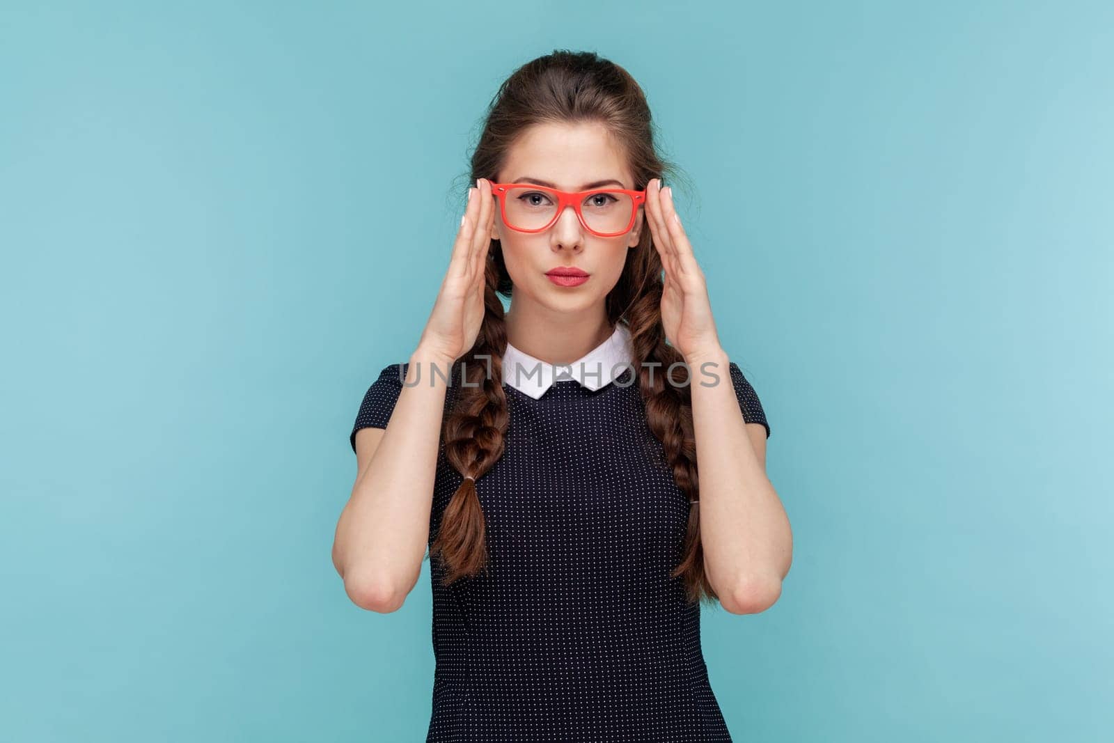 Portrait of serious attractive woman with braids stands keeps hands on frame of red glasses, looking at camera with attention, wearing black dress. woman Indoor studio shot isolated on blue background