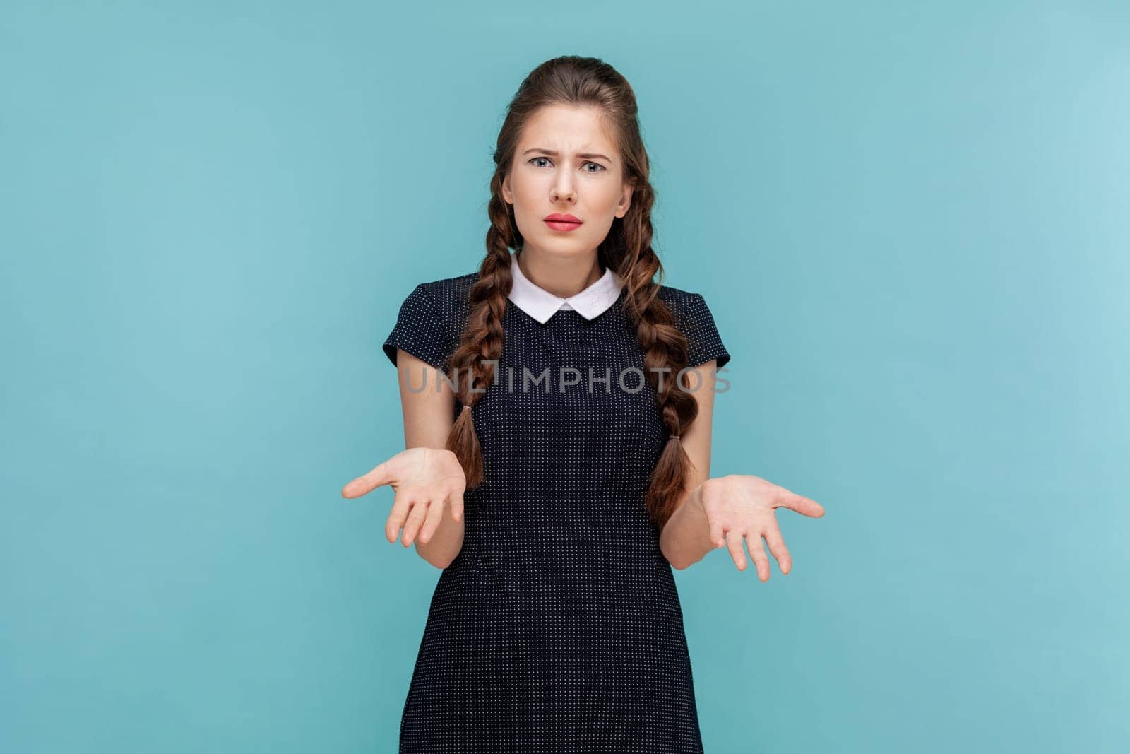 How could you. Portrait of sad annoyed woman with braids standing with spread hands, being irritated, asking what, wearing black dress. woman Indoor studio shot isolated on blue background.