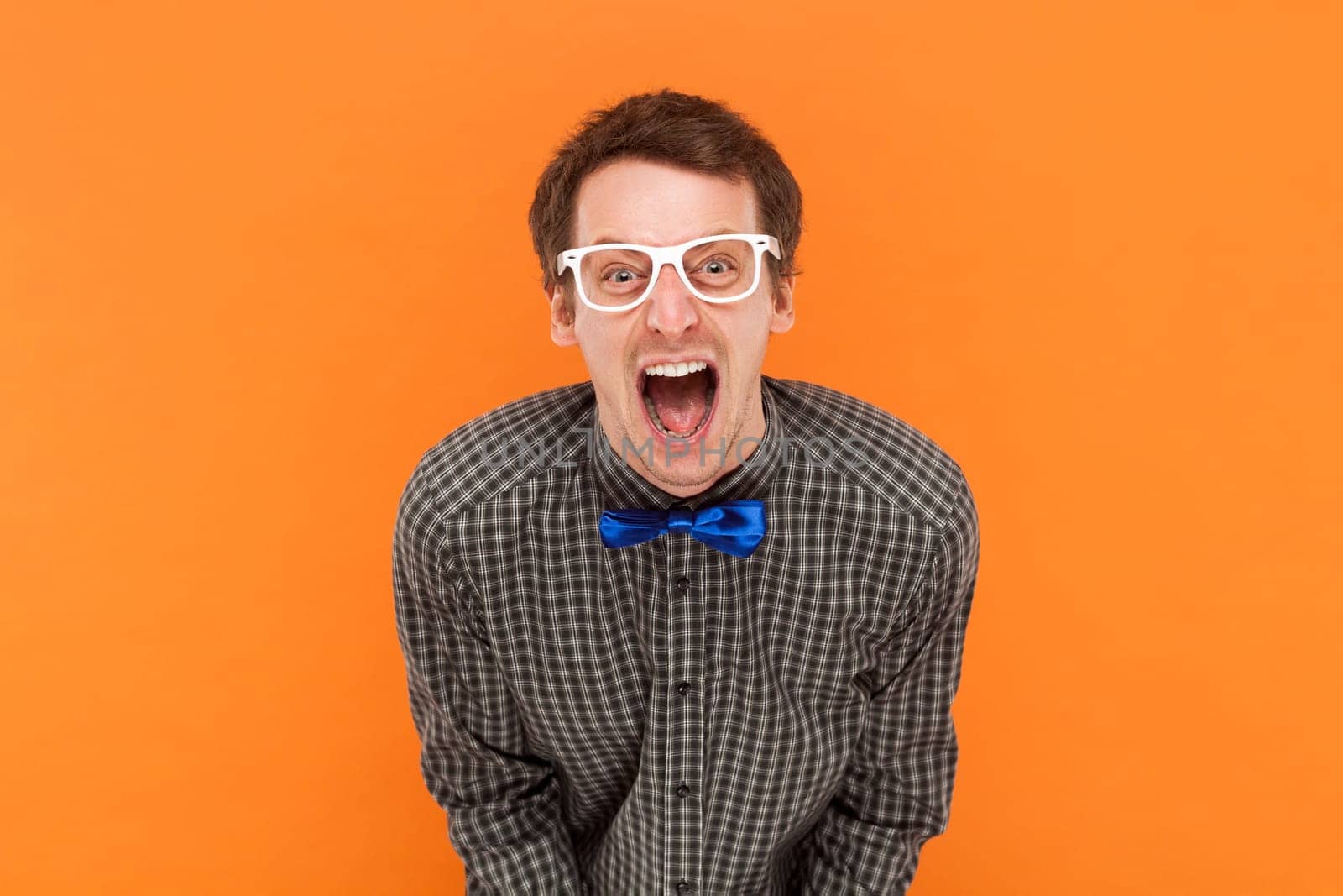 Emotional scared man nerd looks with frightened expression, looks nervously at camera, demonstrates phobia, wears shirt with blue bow tie and glasses. Indoor studio shot isolated on orange background.