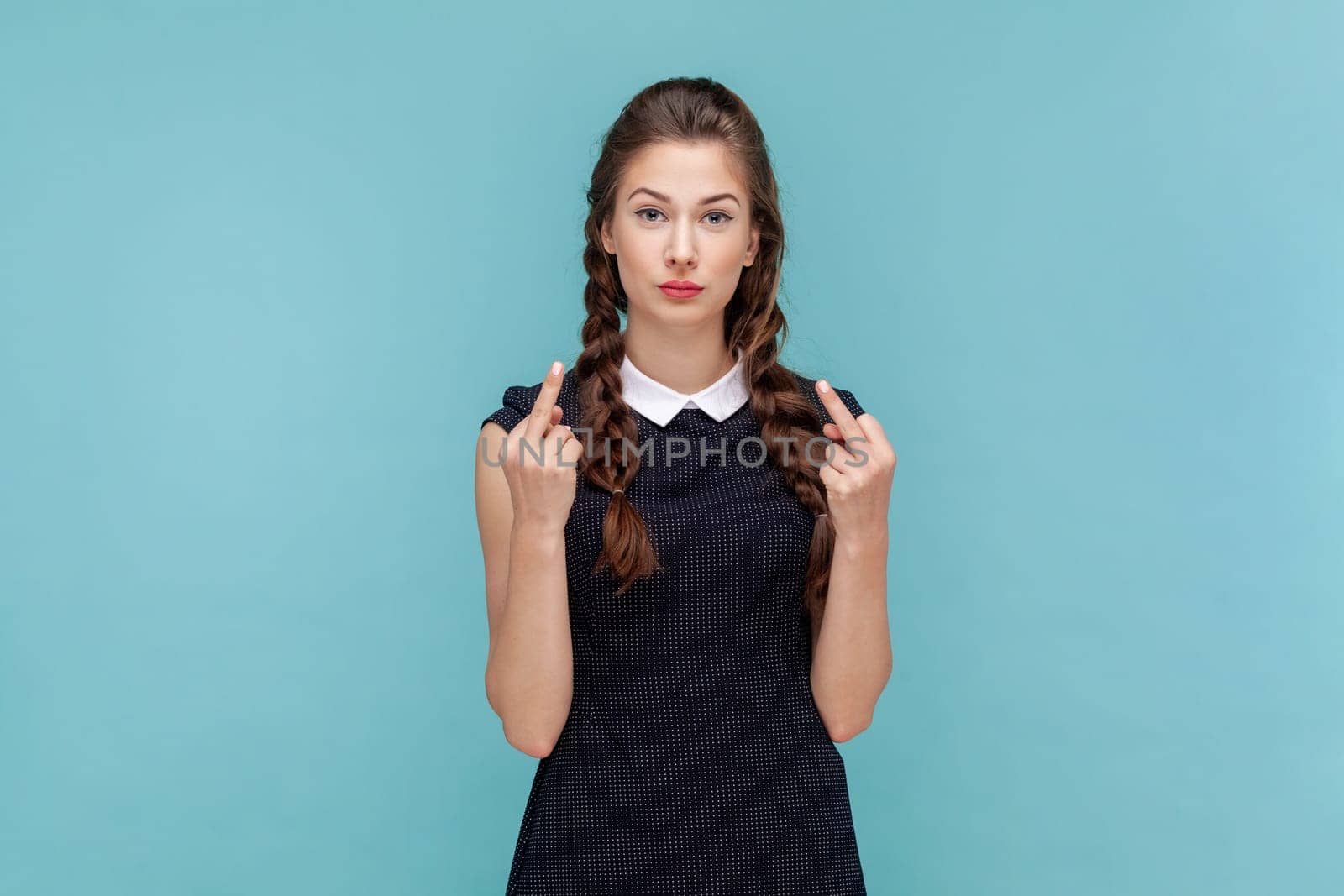 Portrait of serious vulgar woman with braids showing middle finger, impolite gesture disrespects someone looks rude at camera, wearing black dress. woman Indoor studio shot isolated on blue background
