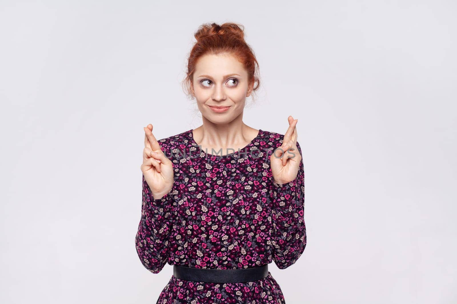 Portrait of redhead woman wearing dress makes wish believes in good luck, hopes dreams come true, has hopeful expression, anticipates for something. Indoor studio shot isolated on gray background.