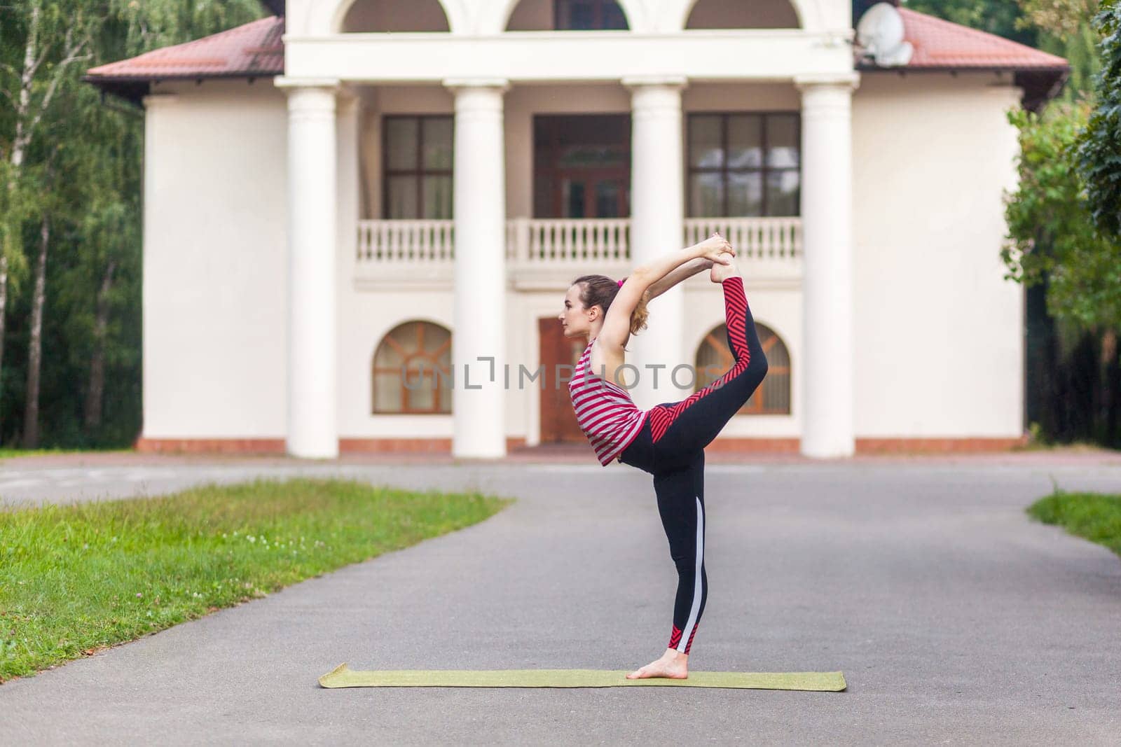 Full length portrait of young attractive woman practicing yoga, standing in natarajasana exercise, holding balance, working out, wearing sportswear, urban style, outdoor sport concept.