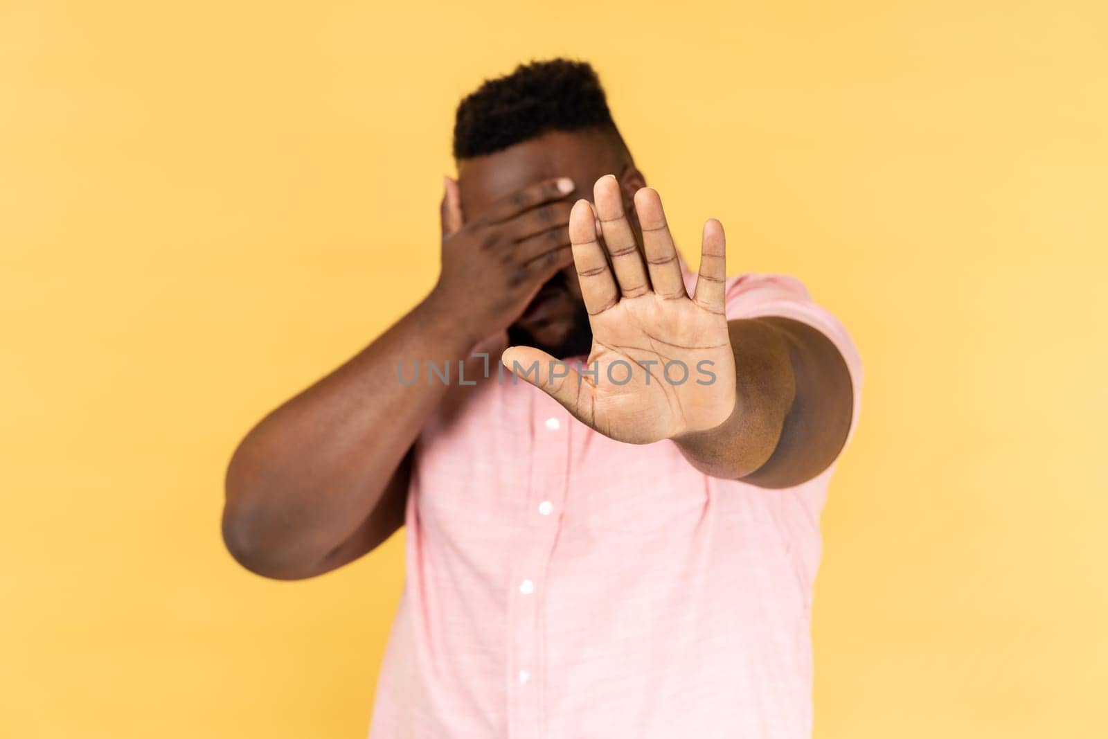 Stop, I don't want to see at this. Portrait of man wearing pink shirt covering his face and showing stop hand gesture. Indoor studio shot isolated on yellow background.
