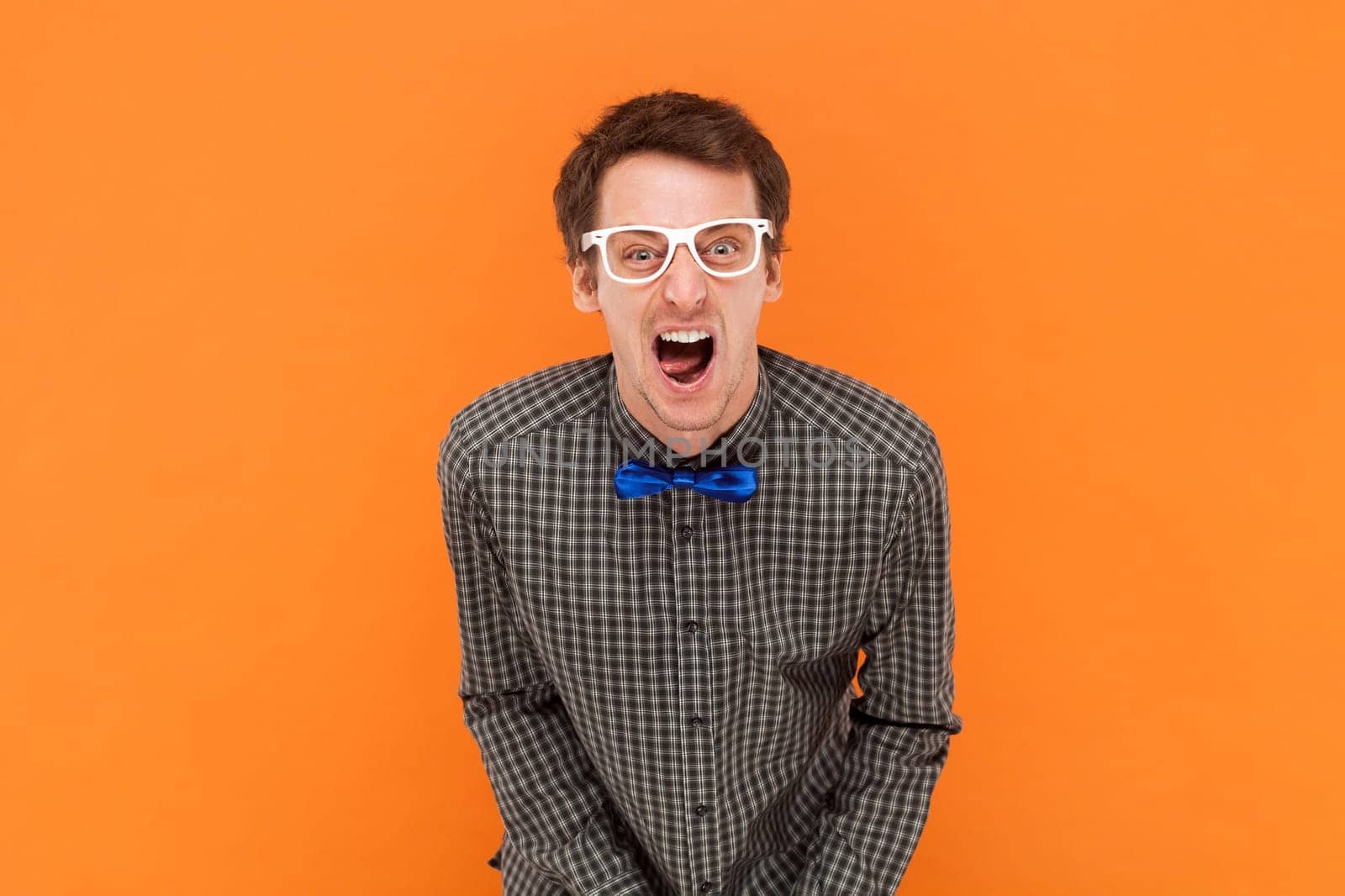 Crazy man nerd yelling loudly, opens mouth broadly, feels desperate after finding out about tragic events, wearing shirt with blue bow tie and glasses. Indoor studio shot isolated on orange background