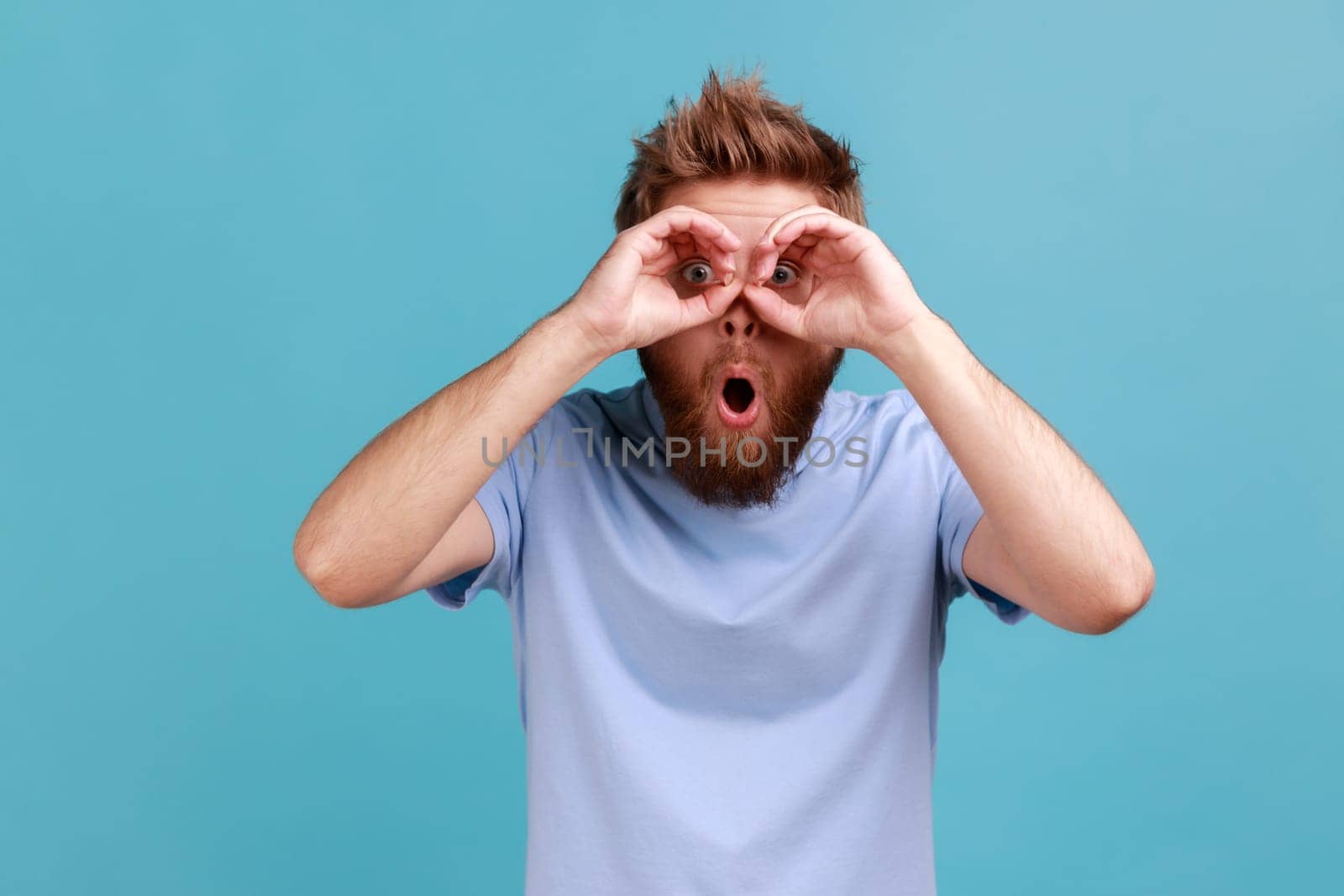 Portrait of wondered shocked bearded man making binoculars gesture with hands and looking amazed at camera, keeps mouth opened. Indoor studio shot isolated on blue background.