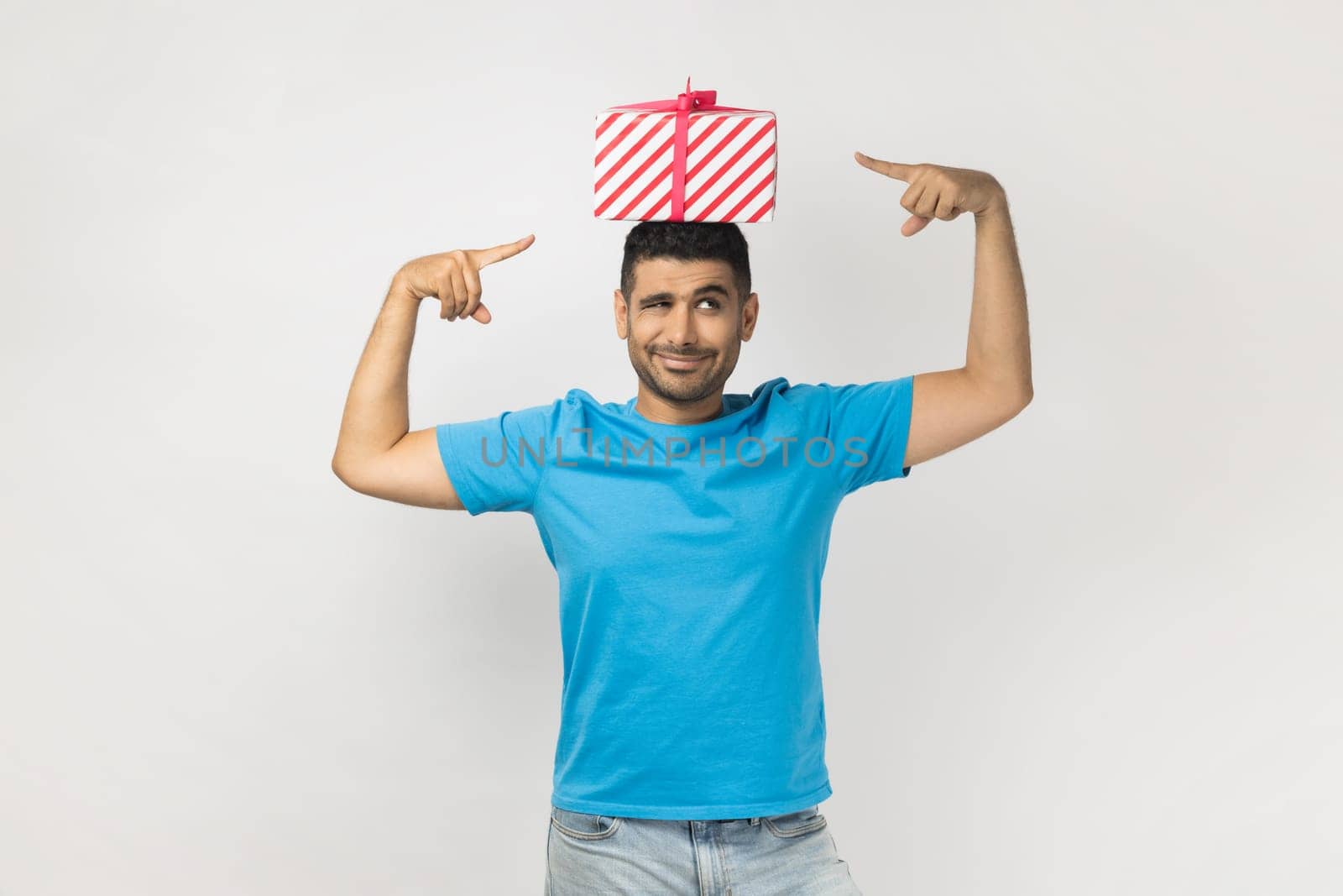 Portrait of optimistic positive unshaven man wearing blue T- shirt standing with gift box on his head, pointing at present, having funny expression. Indoor studio shot isolated on gray background.