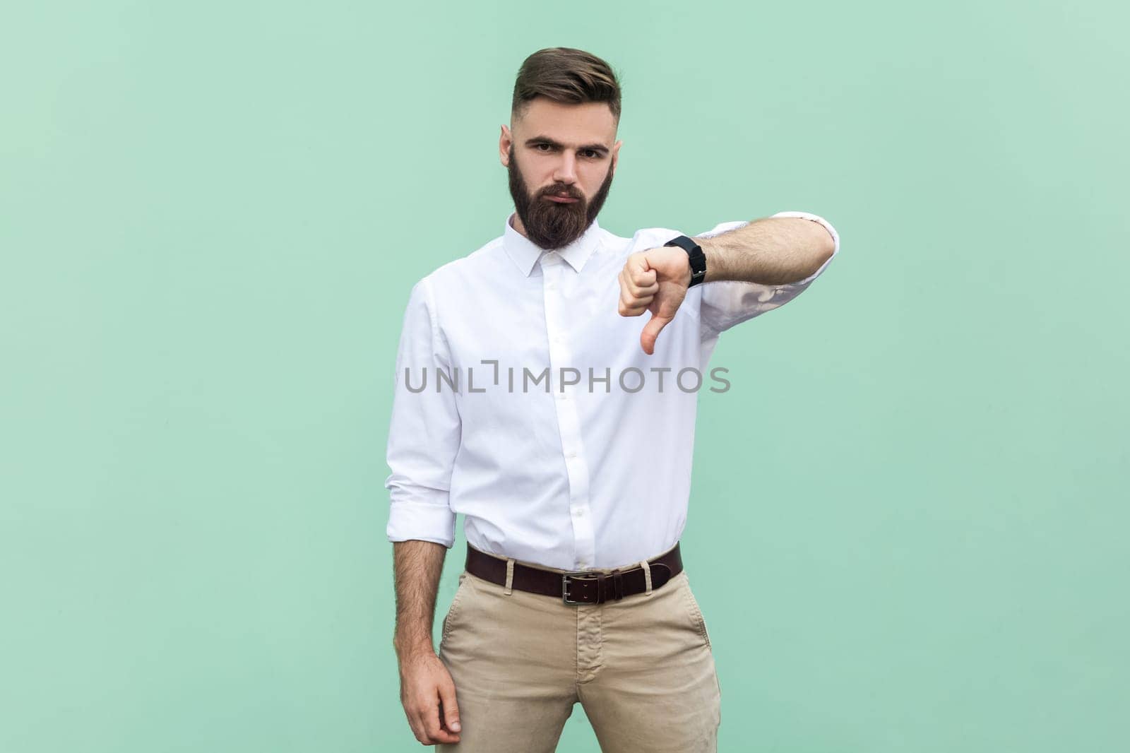 Bearded businessman wearing shirt criticizing bad quality with thumbs down displeased grimace, showing dislike gesture, expressing disapproval. Indoor studio shot isolated on light green background.