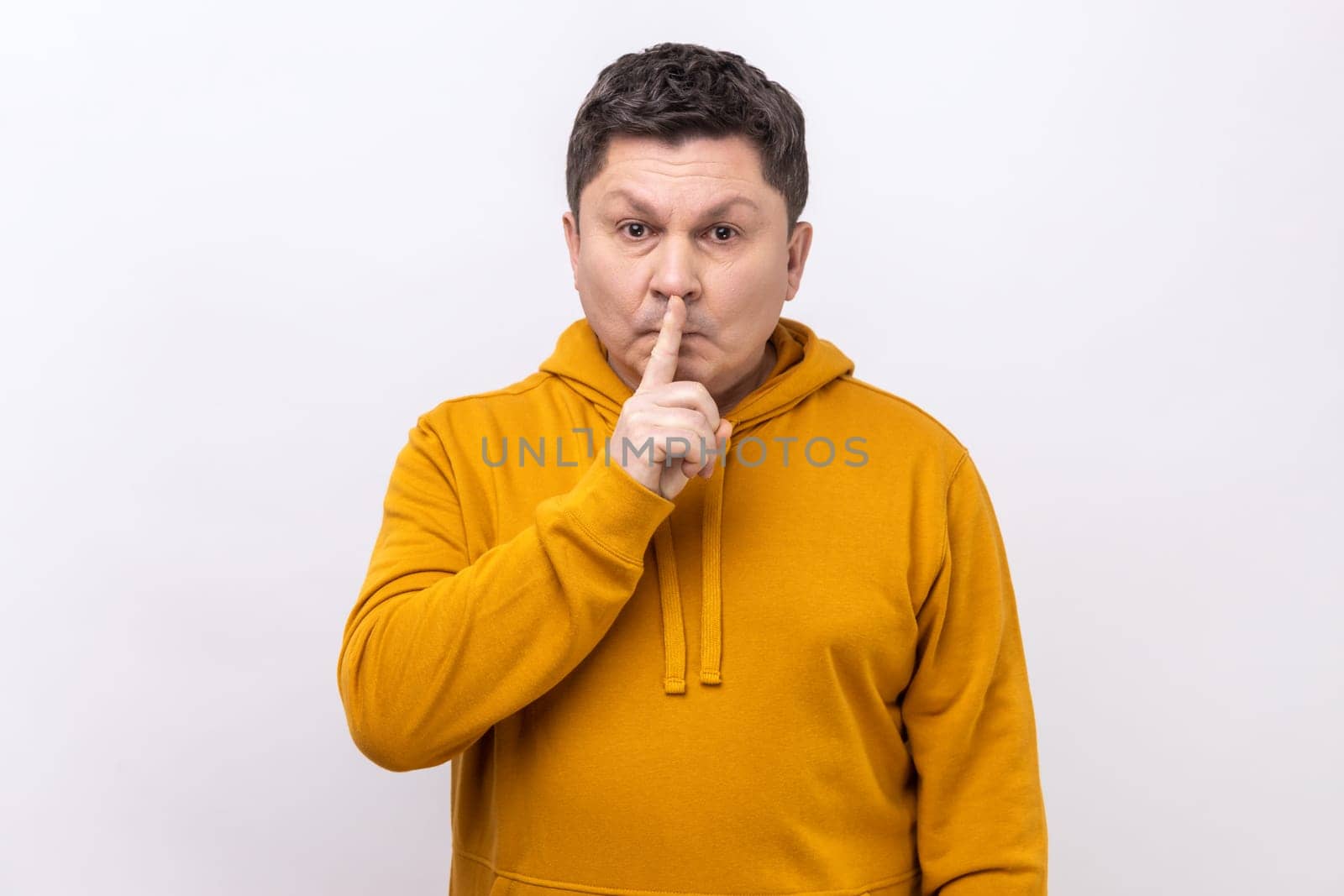 Portrait of middle aged man holding finger on lips making hush silence gesture, asking to keep secret, don't speak, wearing urban style hoodie. Indoor studio shot isolated on white background.