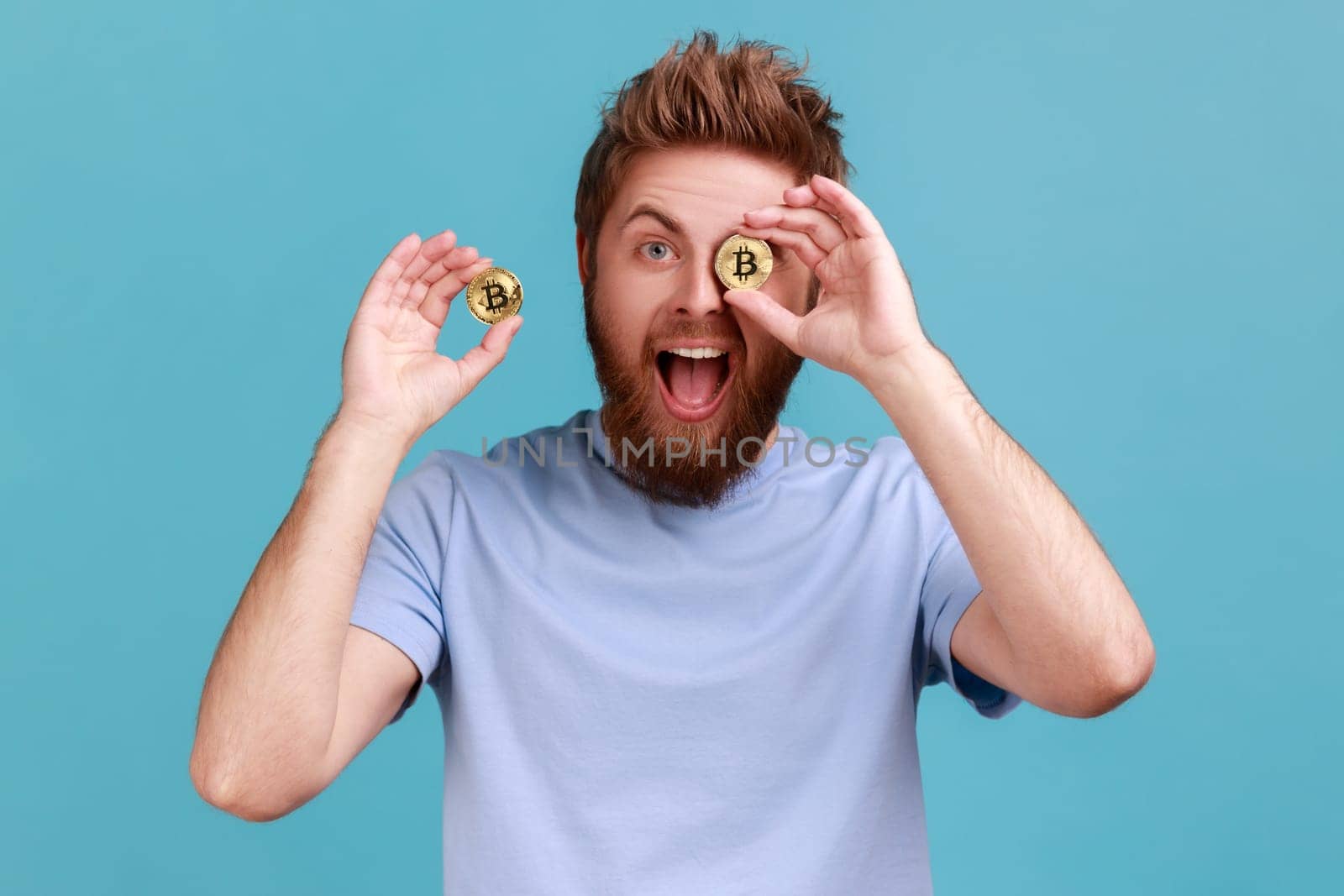 Portrait of excited positive bearded man with open mouth covering eye with golden btc coin and smiling to camera, advertising of cryptocurrency. Indoor studio shot isolated on blue background.