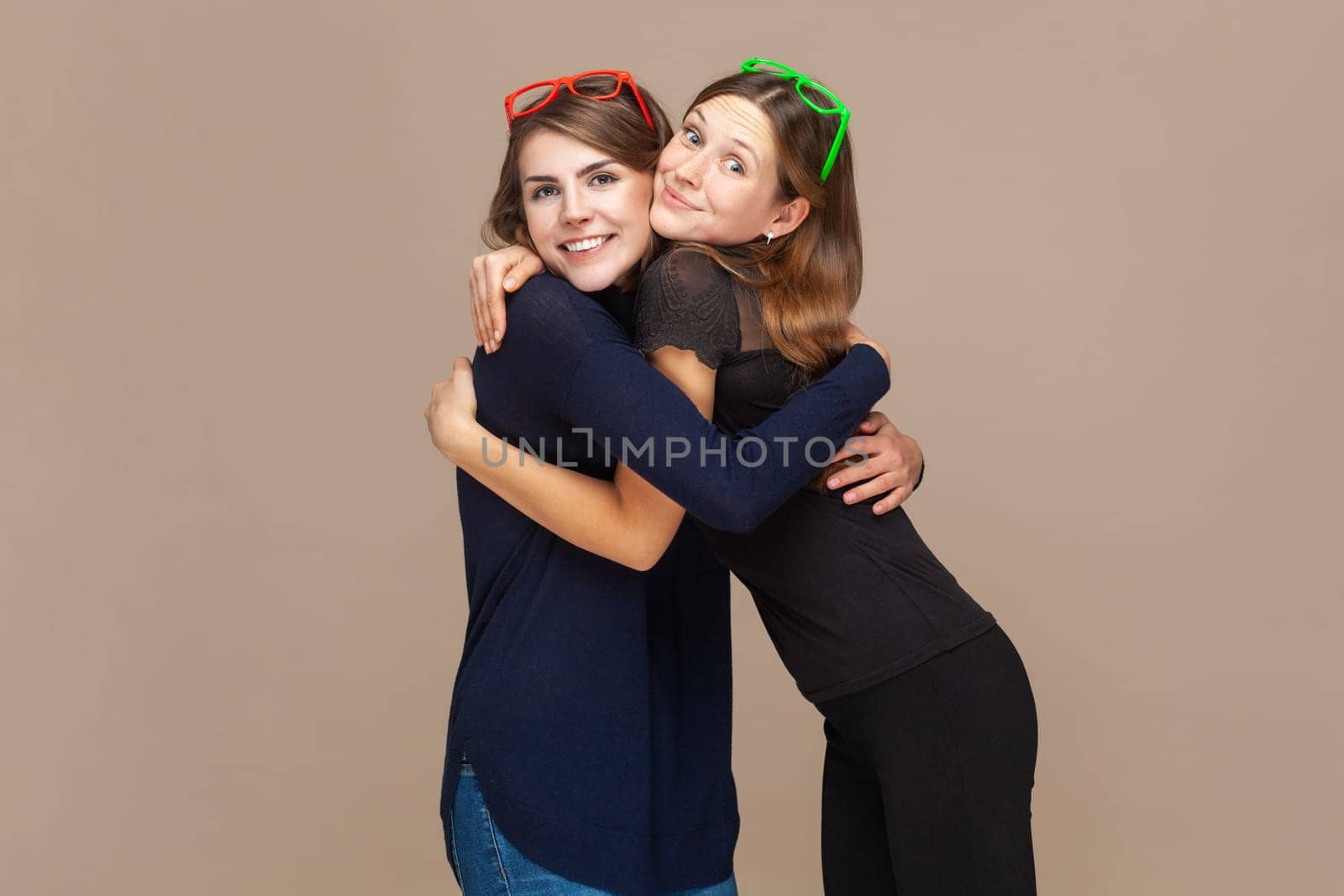 Portrait of two smiling happy women best friends sanding hugging each other, looking at camera, expressing positive. Indoor studio shot isolated on light brown background.