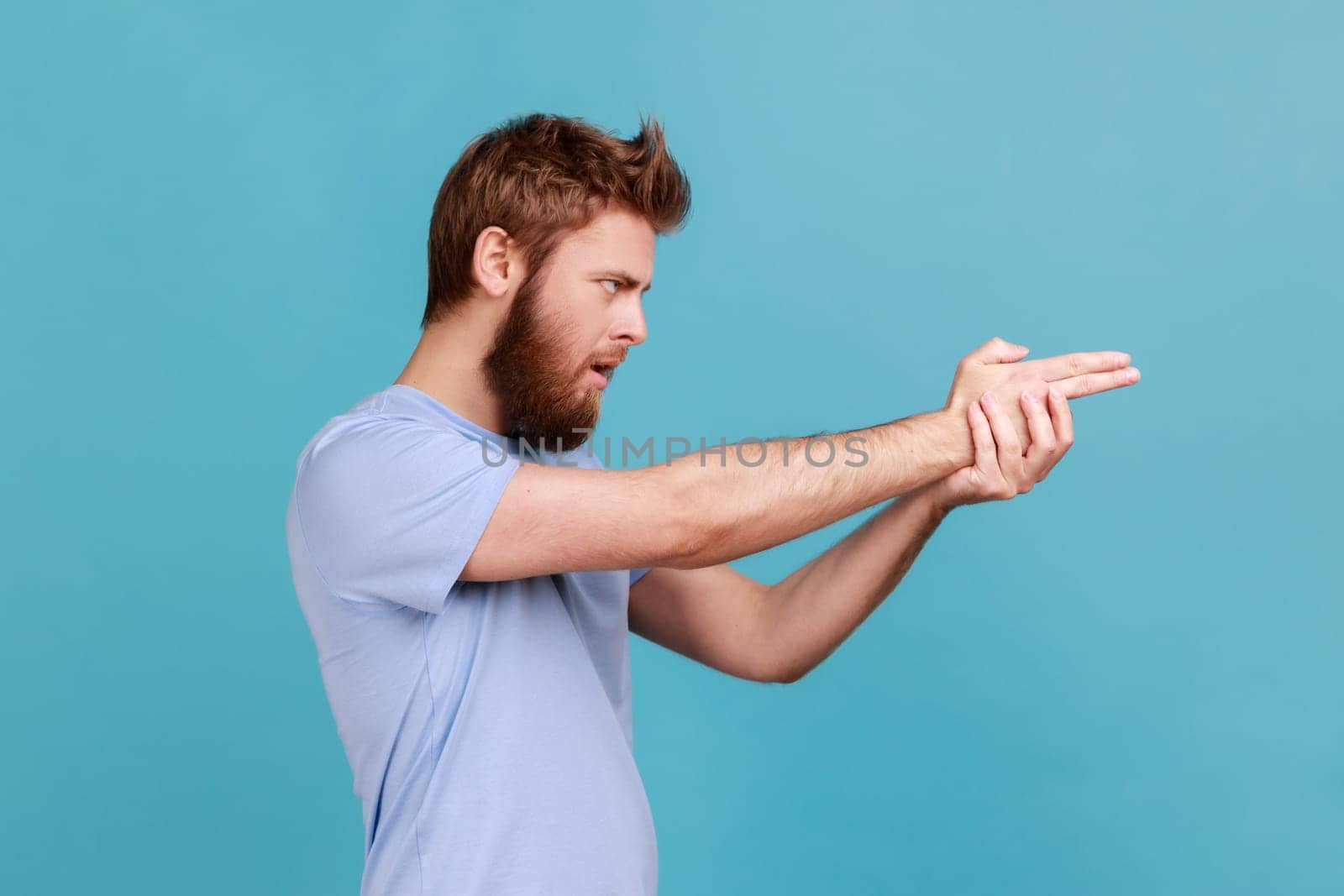 Side view of focused bearded man pointing finger gun gesture to target, threatening to kill, shooting with hand pistol and looking menace. Indoor studio shot isolated on blue background.