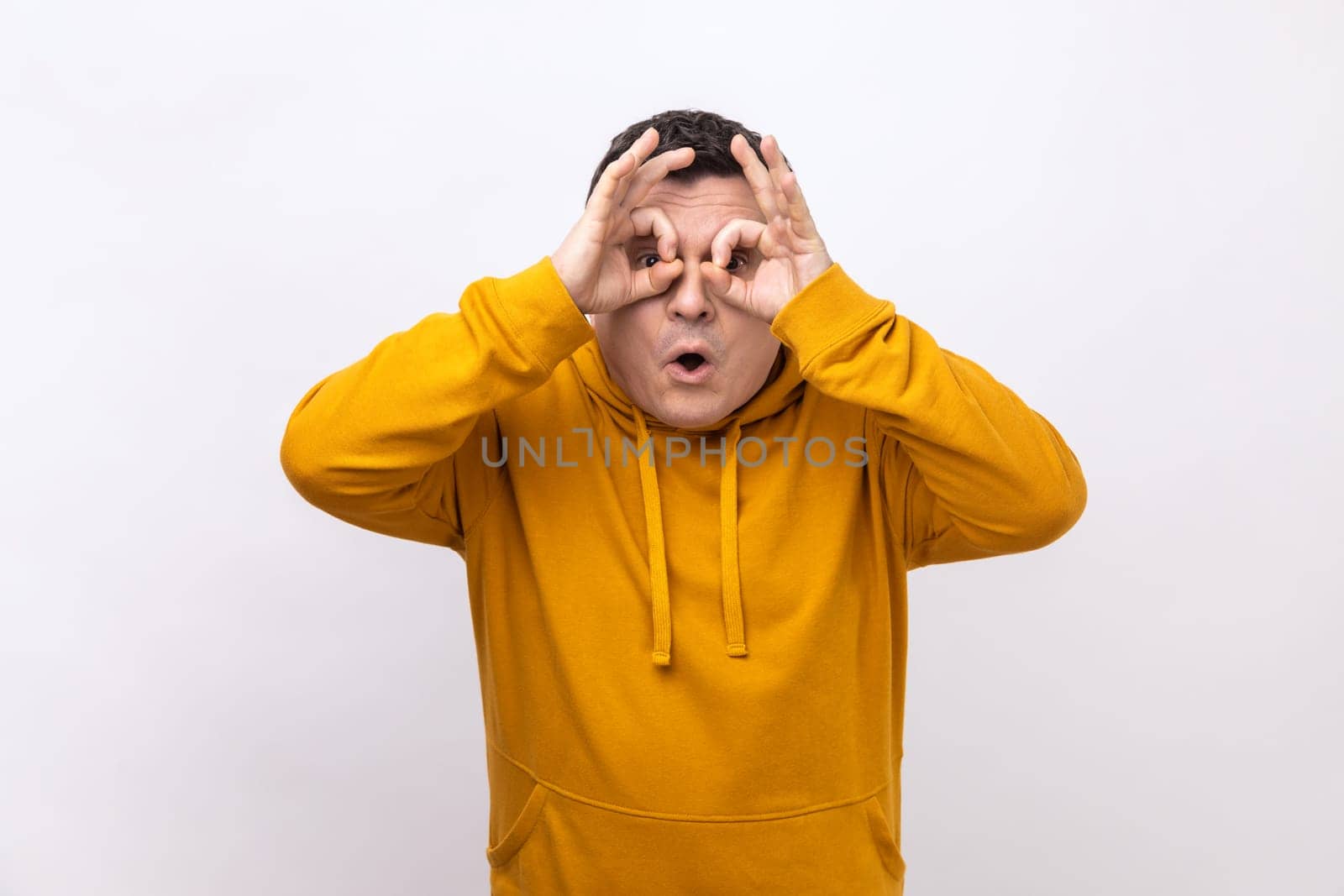 Portrait of curious man looks through fingers in binoculars gesture, observing distant with astonished look, watching afar, wearing urban style hoodie. Indoor studio shot isolated on white background.