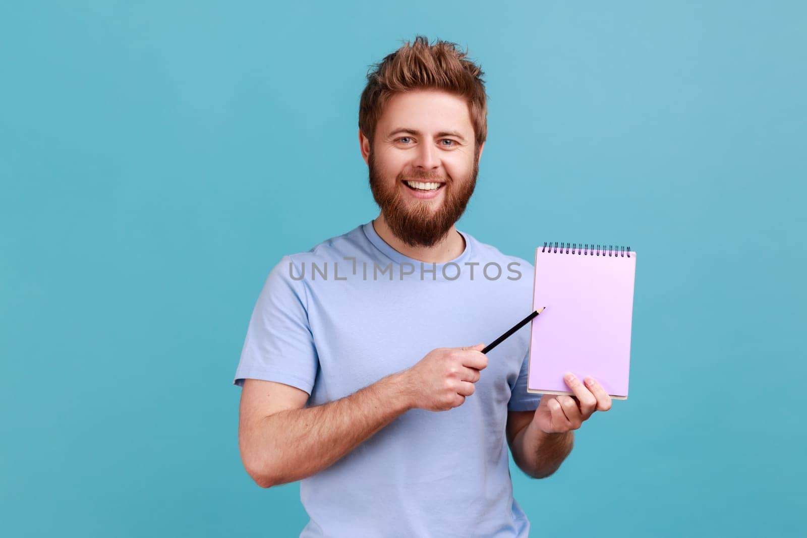 Portrait of bearded man pointing empty sheet and smiling at camera, showing paper notebook template, mock up blank space for business idea, message. Indoor studio shot isolated on blue background.