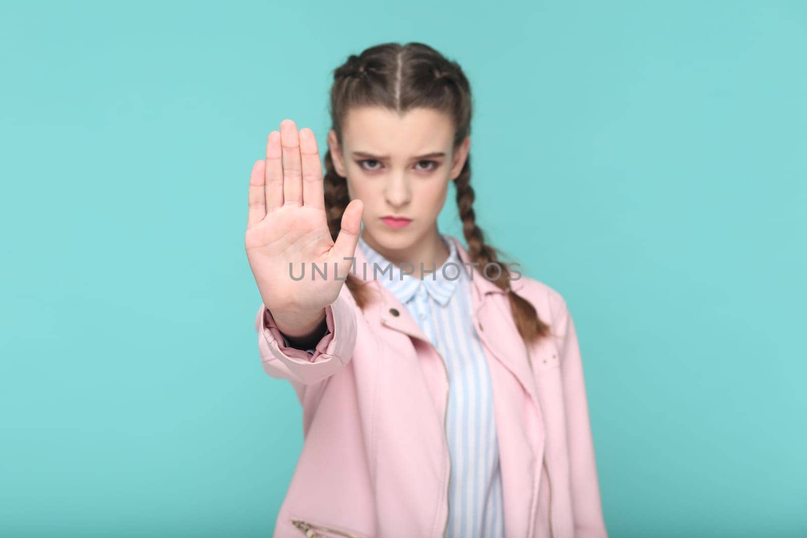 Portrait of serious strict teenager girl with braids wearing pink jacket showing stop gesture, looking at camera with angry look. Indoor studio shot isolated on green background.