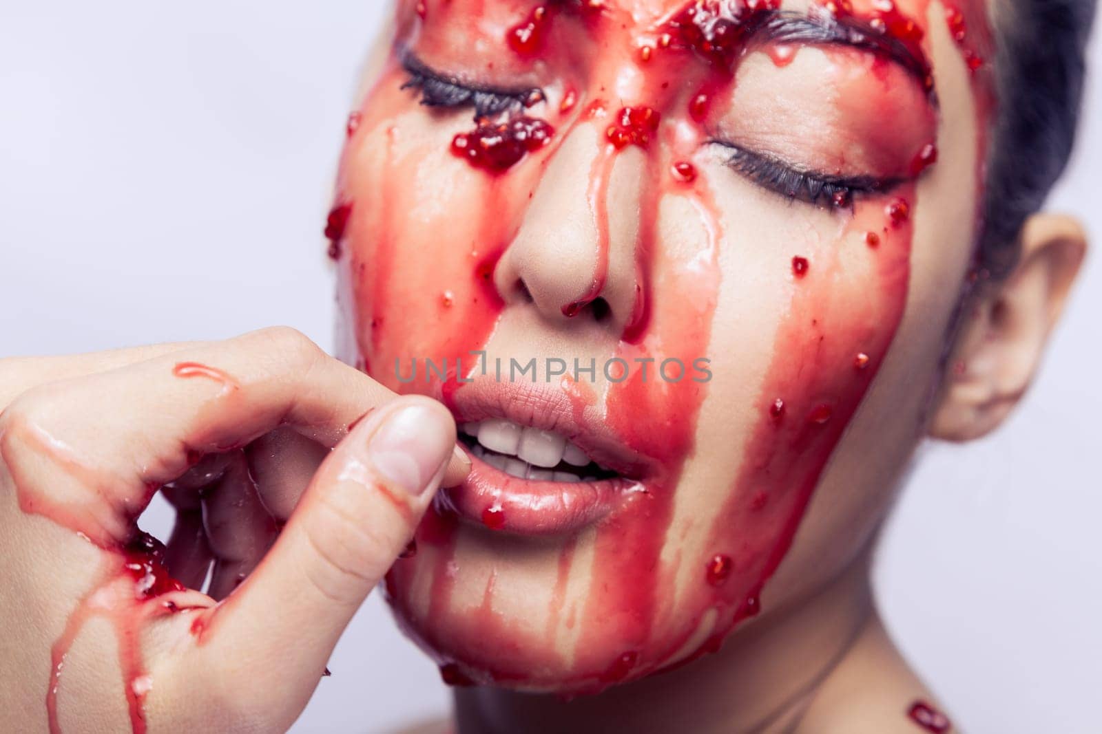 Closeup portrait of woman model with open mouth and closed eyes touching fingers her lips and tries, jam flowing on her face. Indoor studio shot isolated on gray background.