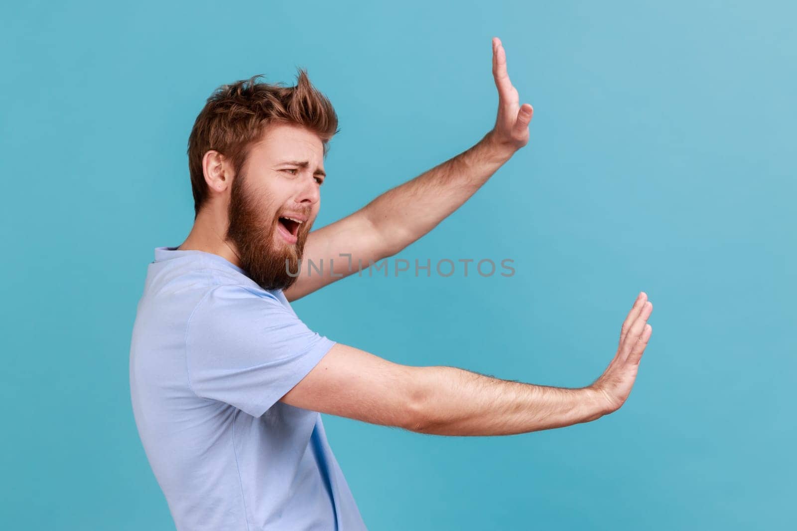Side view of shocked scared unhappy young bearded man screaming in horror and fear, keeping hands raised to defend himself, panic attack. Indoor studio shot isolated on blue background.