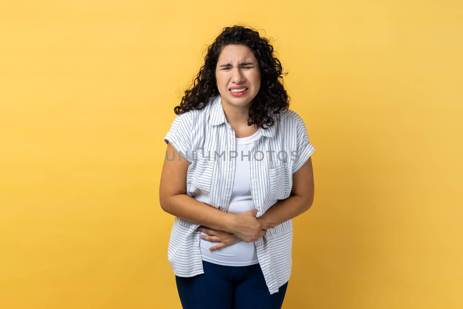Portrait of sad depressed unhealthy woman with dark wavy hair suffering from strong stomachache, frowning, pain during period. Indoor studio shot isolated on yellow background.