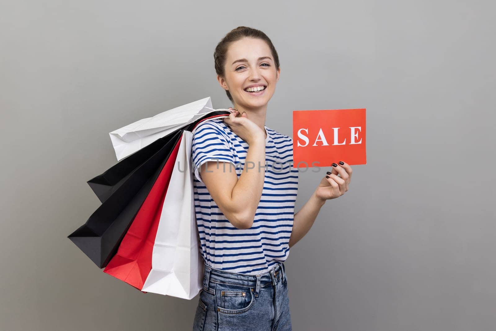Side view of delighted woman wearing striped T-shirt boasting with purchases holding shopping bags and sale card, big holidays discounts. Indoor studio shot isolated on gray background.