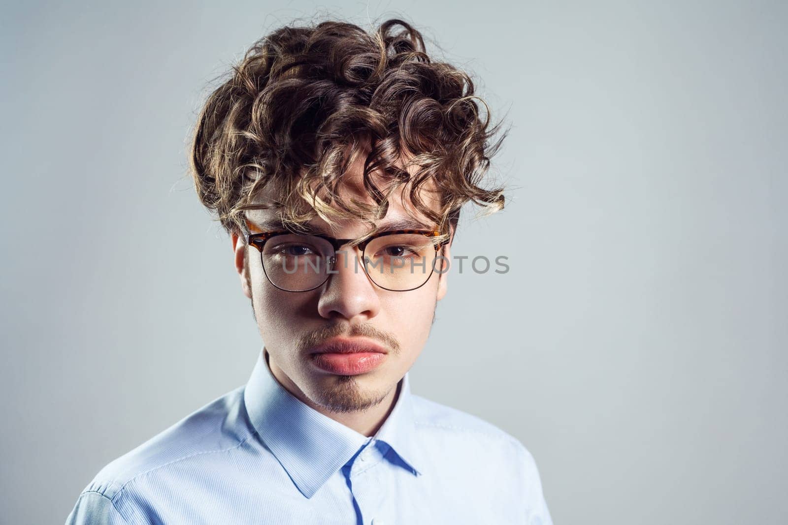 Portrait of serious young adult man wearing blue shirt and eyeglasses, with curly hairstyle, looking at camera with strict expression. Indoor studio shot isolated on gray background.