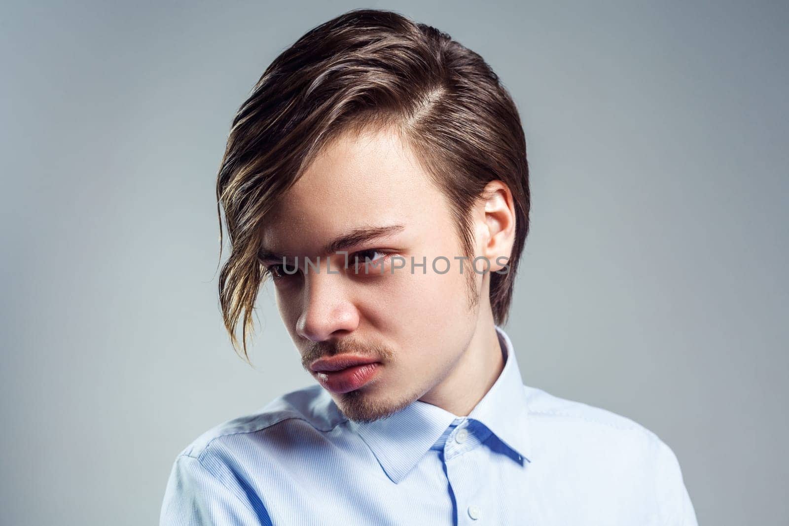 Portrait of young attractive man with long fringe hairstyle on his eyes, looking away with mysterious look, wearing blue shirt. Indoor studio shot isolated on gray background.