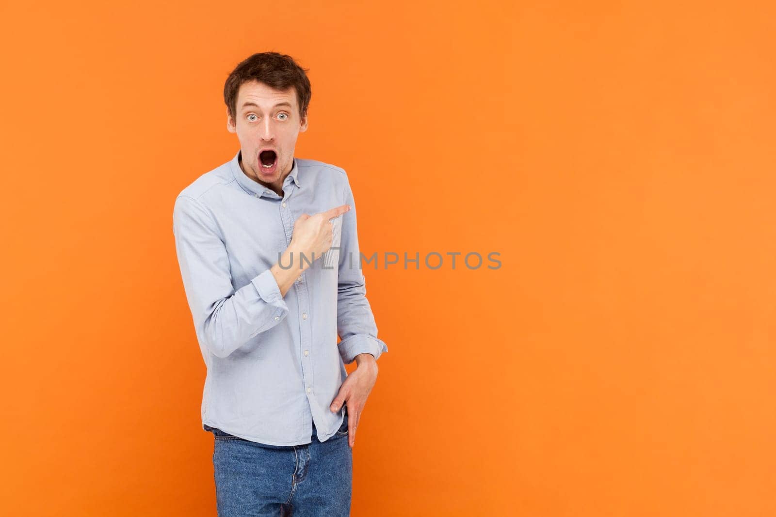 Shocked amazed young adult man pointing aside with finger, showing copy space for advertisement or promotional text, wearing light blue shirt. Indoor studio shot isolated on orange background.
