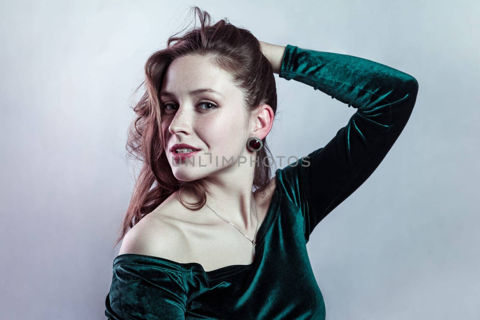 Beauty portrait of smiling cunning attractive woman wearing green dress keeps hand on her hair, looking at camera confident expression. Indoor studio shot isolated on gray background.