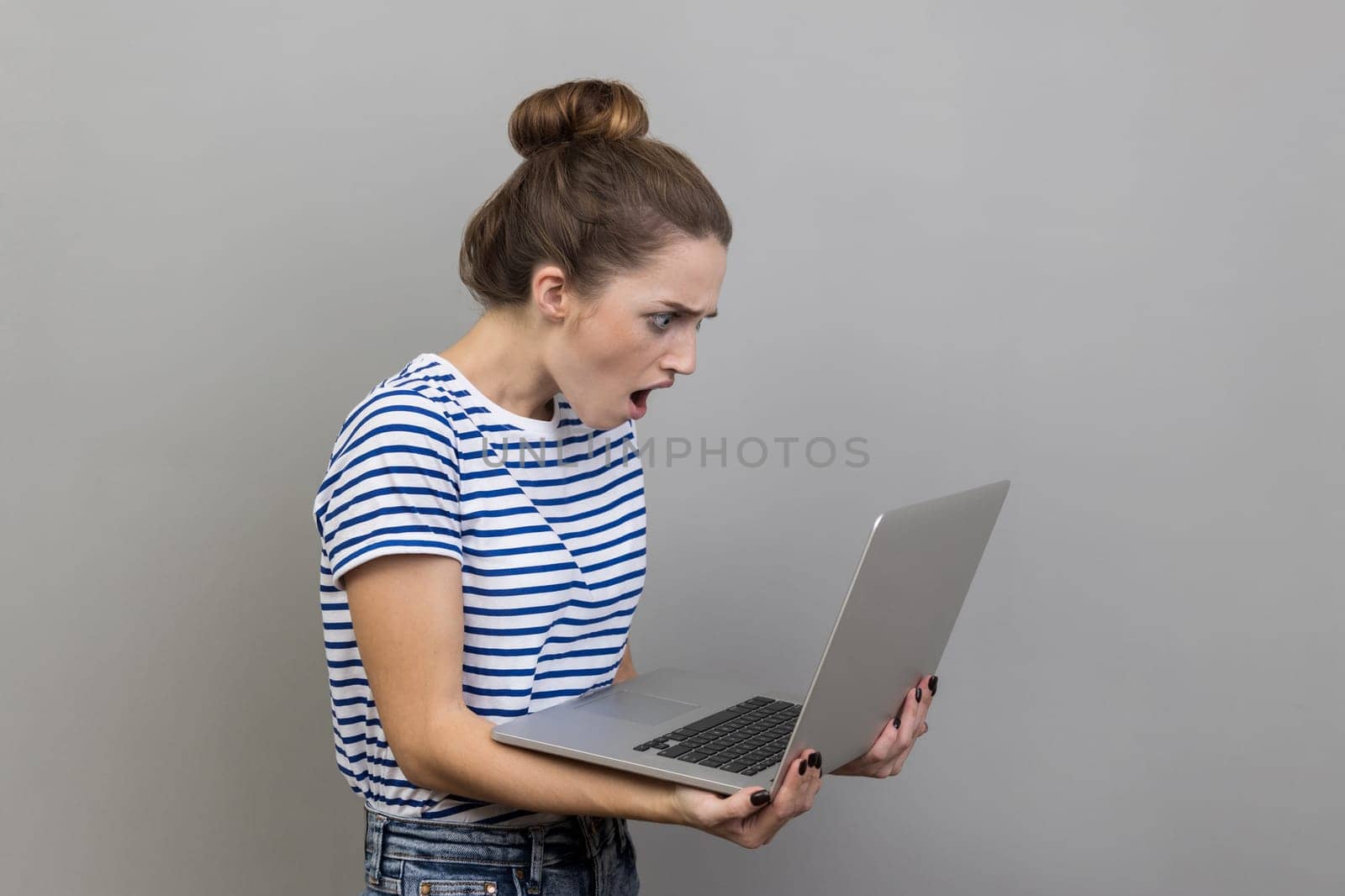 Side view of shocked woman wearing striped T-shirt working on laptop with surprised expression, freelance work, looking at display. Indoor studio shot isolated on gray background.