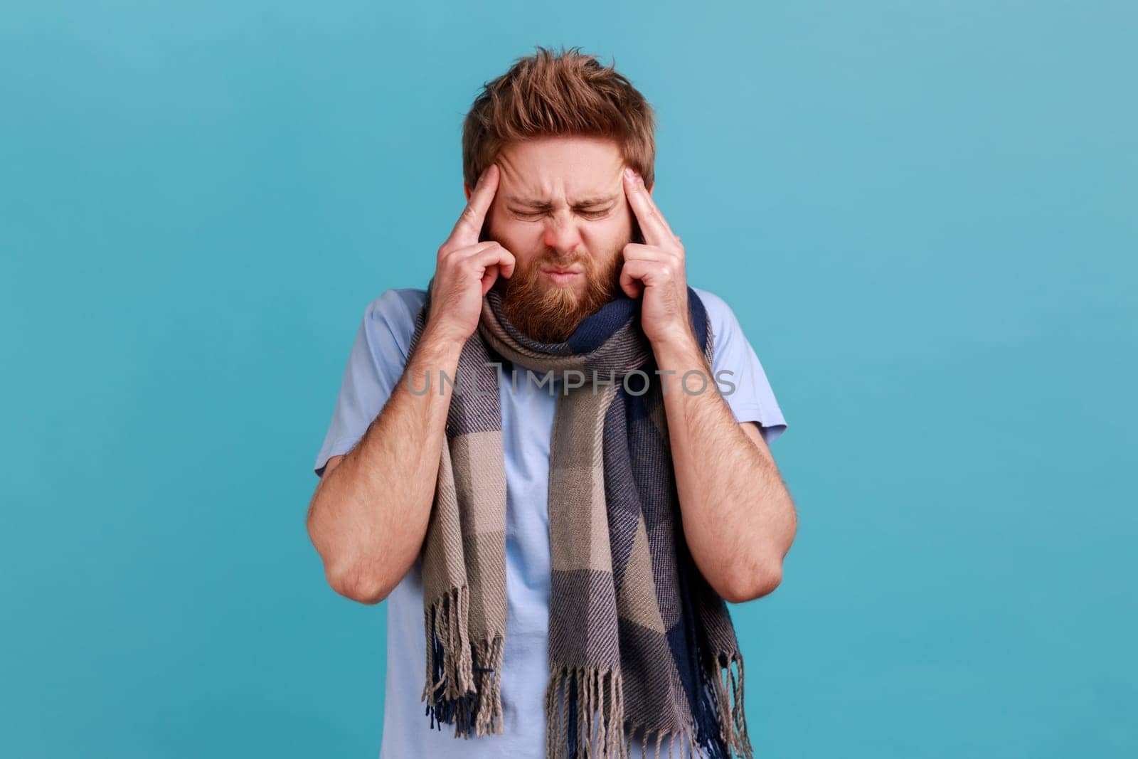 Portrait of bearded man wrapped in scarf standing with painful grimace, touching sore head, suffering migraine, sick with influenza. Indoor studio shot isolated on blue background.