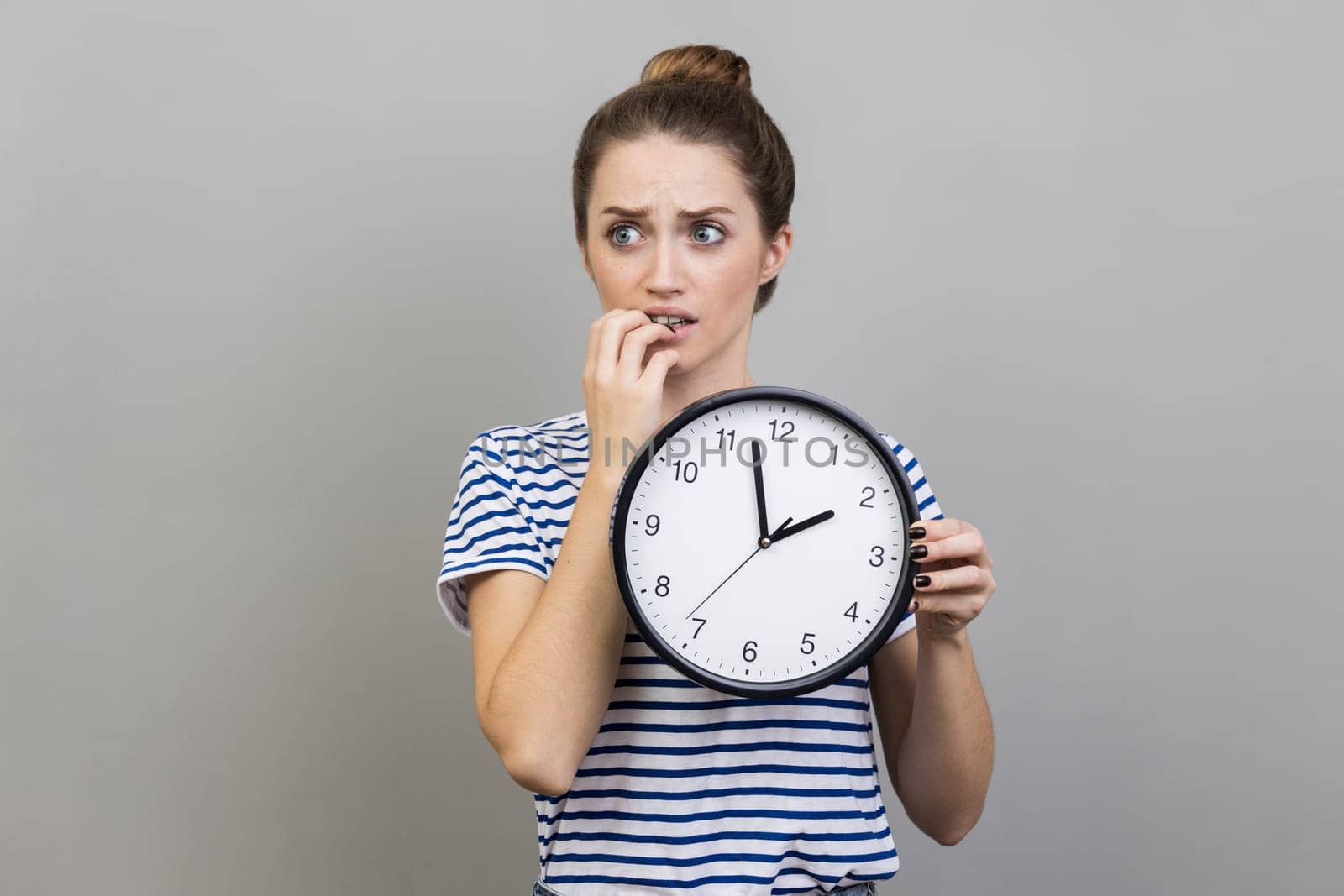 Portrait of woman wearing striped T-shirt holding wall clock in hands, biting nails, looking at camera with nervous expression, deadline. Indoor studio shot isolated on gray background.