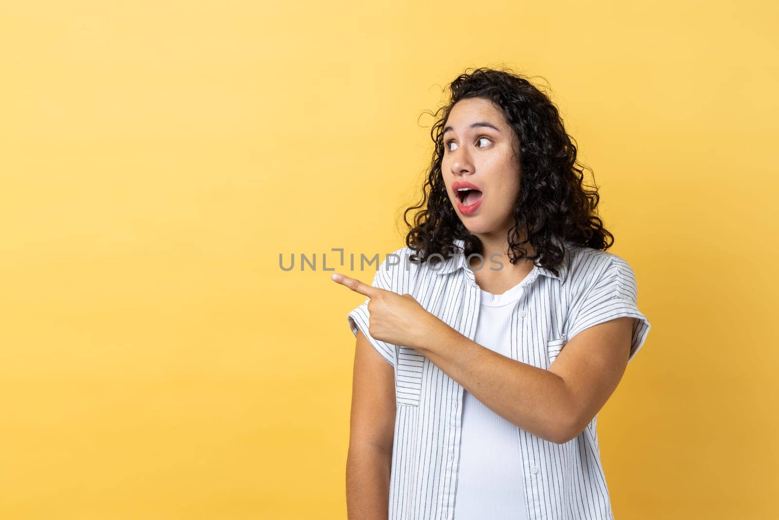 Portrait of shocked astonished attractive woman with dark wavy hair pointing and looking aside, mockup for promotion or advertisement. Indoor studio shot isolated on yellow background.