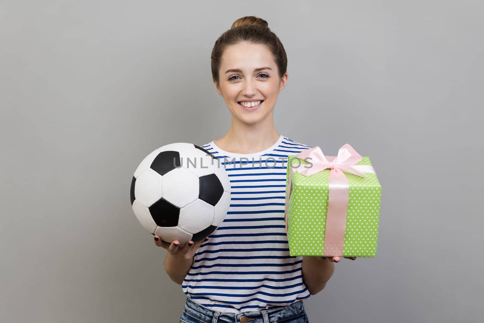 Portrait of woman wearing striped T-shirt standing looking smiling at camera, holding out soccer ball and green present box. Indoor studio shot isolated on gray background.