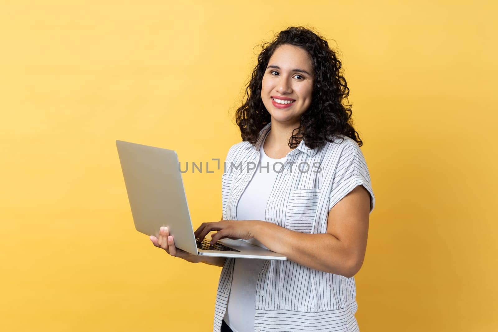 Portrait of satisfied positive happy woman with dark wavy hair working on laptop, looking at camera screen with pleasant expression. Indoor studio shot isolated on yellow background.