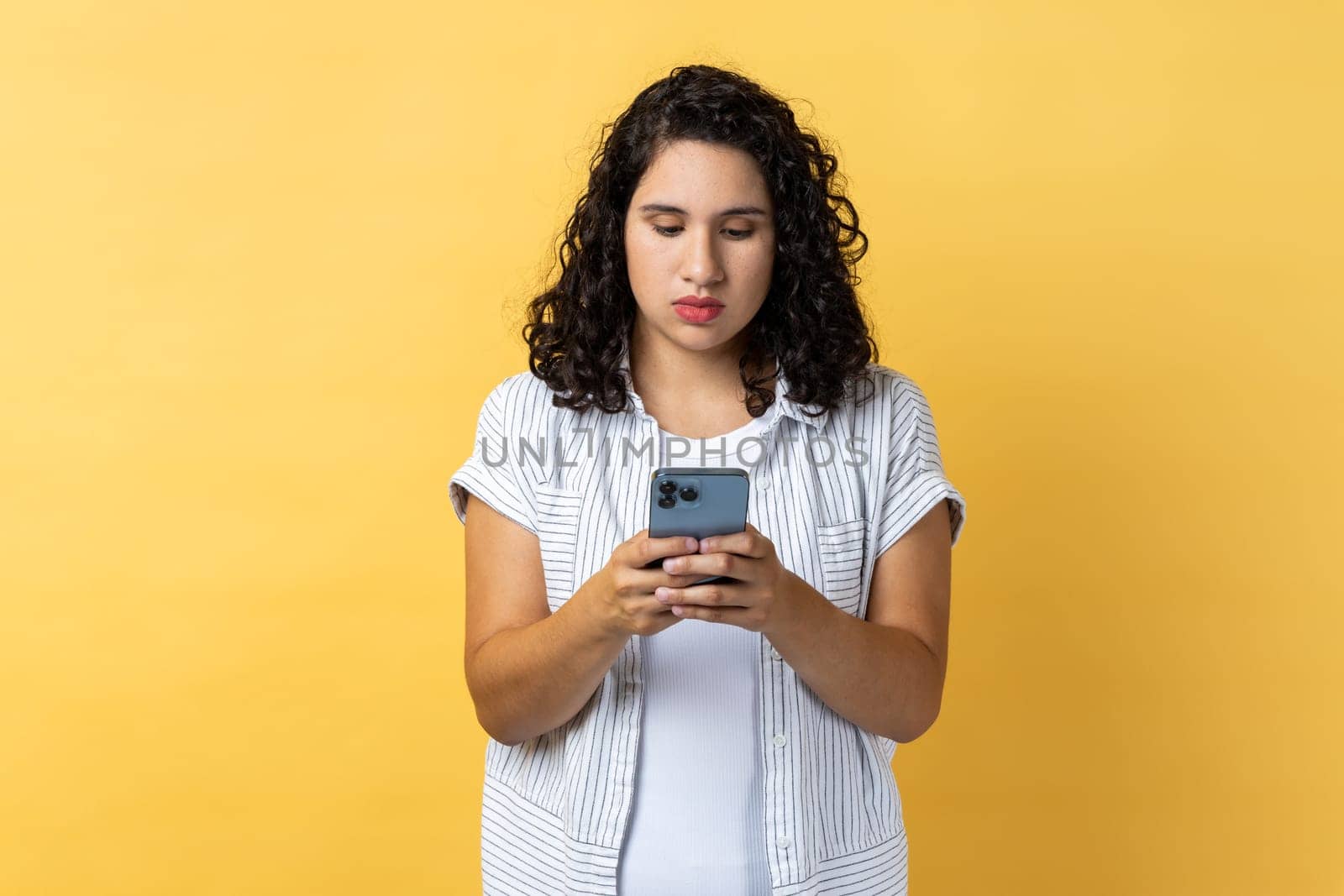 Portrait of serious strict woman with dark wavy hair using smart phone, typing, answering messages, chatting in social networks. Indoor studio shot isolated on yellow background.