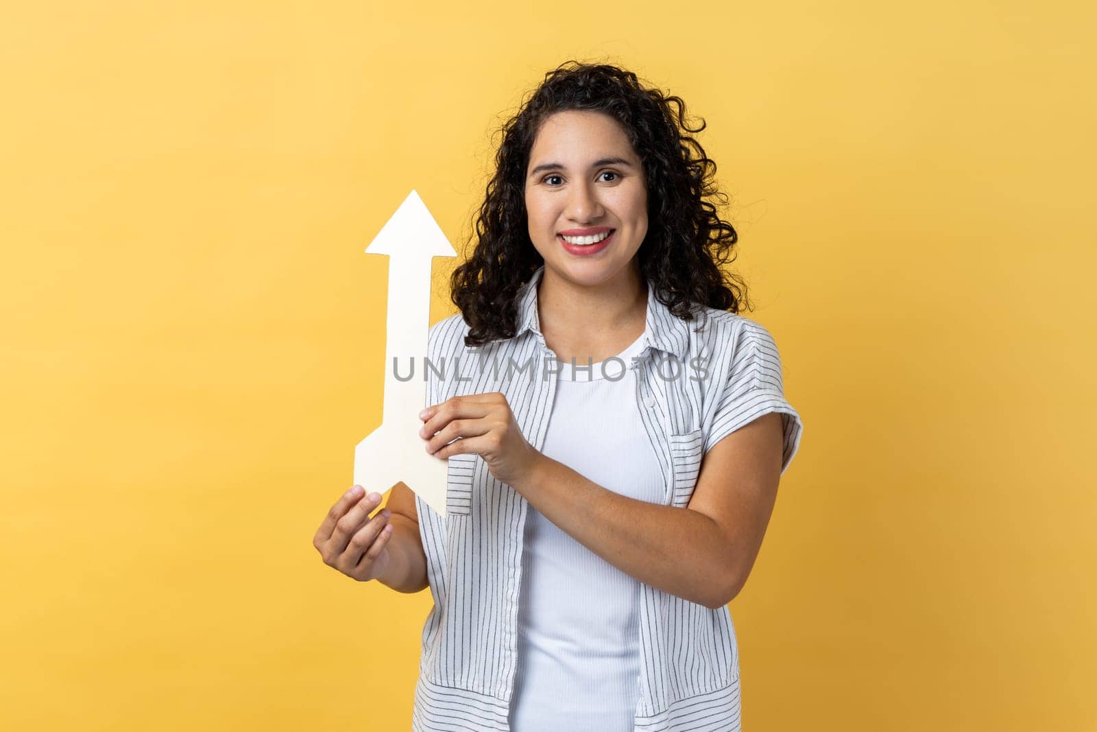 Portrait of smiling satisfied woman with dark wavy hair pointing up looking at camera with smile, growth and increase concept. Indoor studio shot isolated on yellow background.